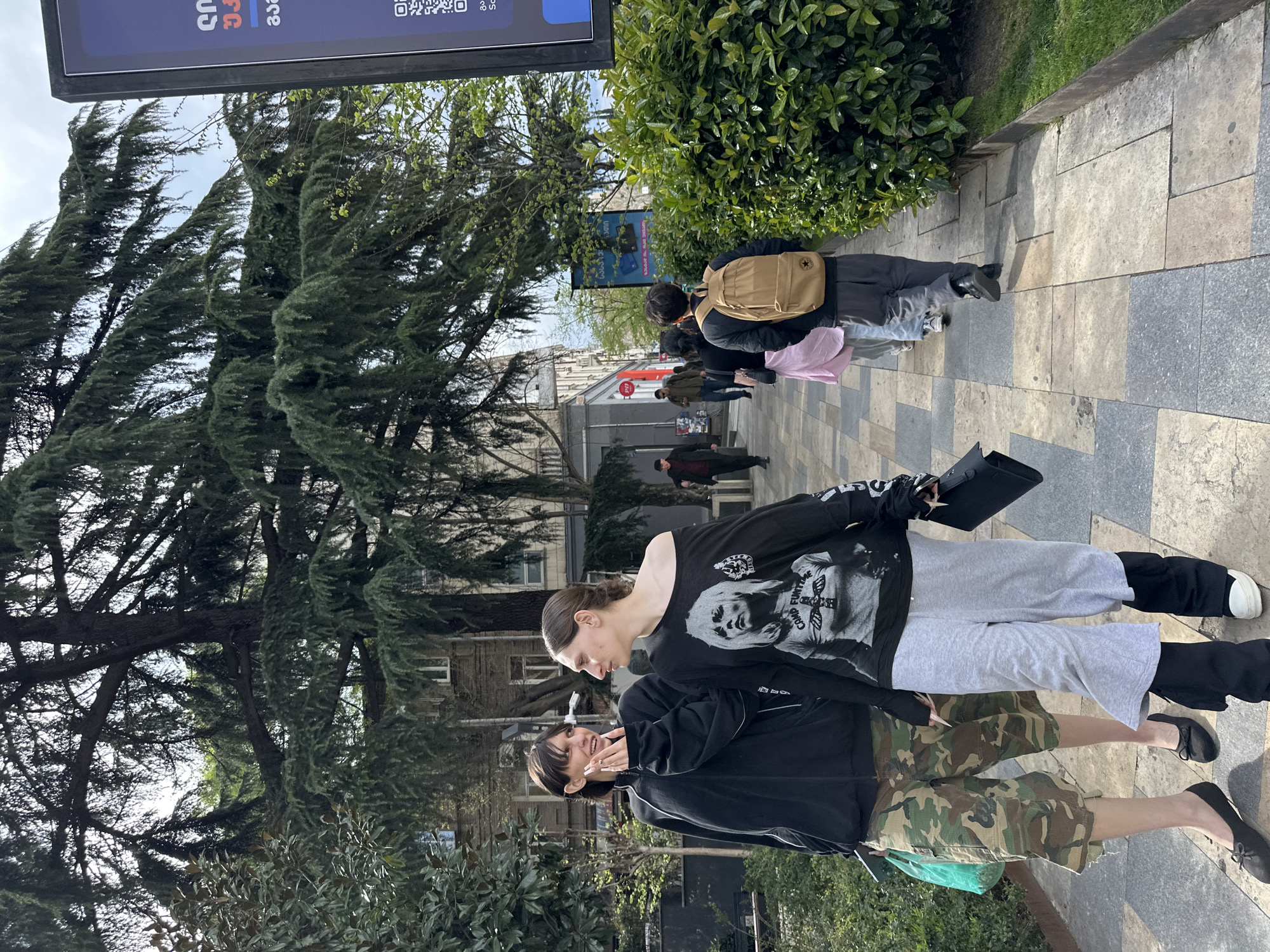A daytime street scene in Tbilisi, Georgia. A tree dominates the left side of the image. The right side shows a sidewalk with pedestrians walking in both directions. In the foreground, two individuals are conversing. One is wearing a black long-sleeve shirt with a graphic design, grey sweatpants, and black shoes. The other individual is wearing a black jacket and camouflage shorts. In the background, people are walking on the sidewalk with a building and a poster.
