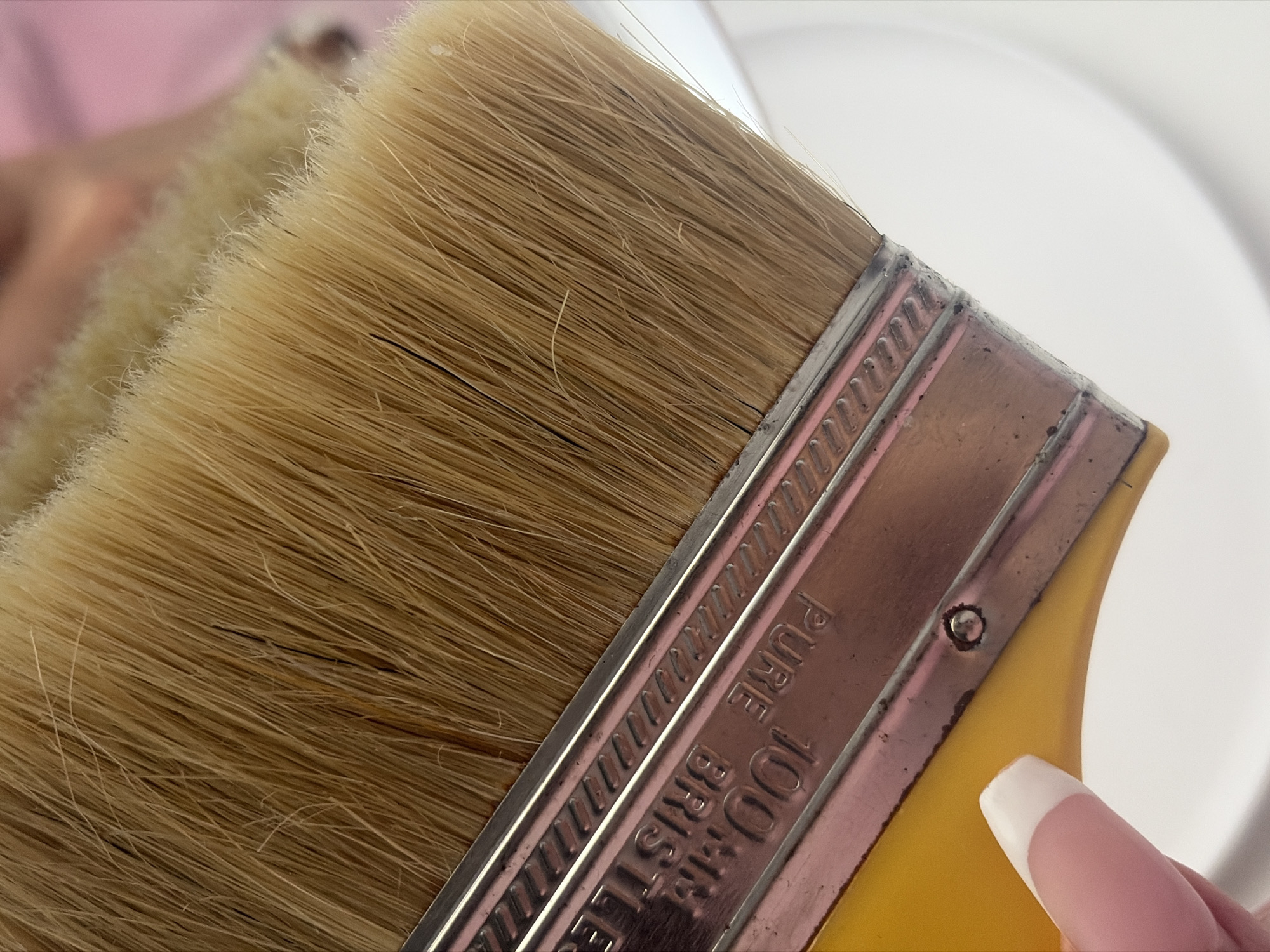 A close-up image shows a tan-colored paintbrush with light bristles. The brush has a metal band with the words "PURE 100% BRISTLE" etched on it. The handle appears to be yellow. A finger with a French manicure is visible near the handle. The background is blurred, with hints of white and pink.