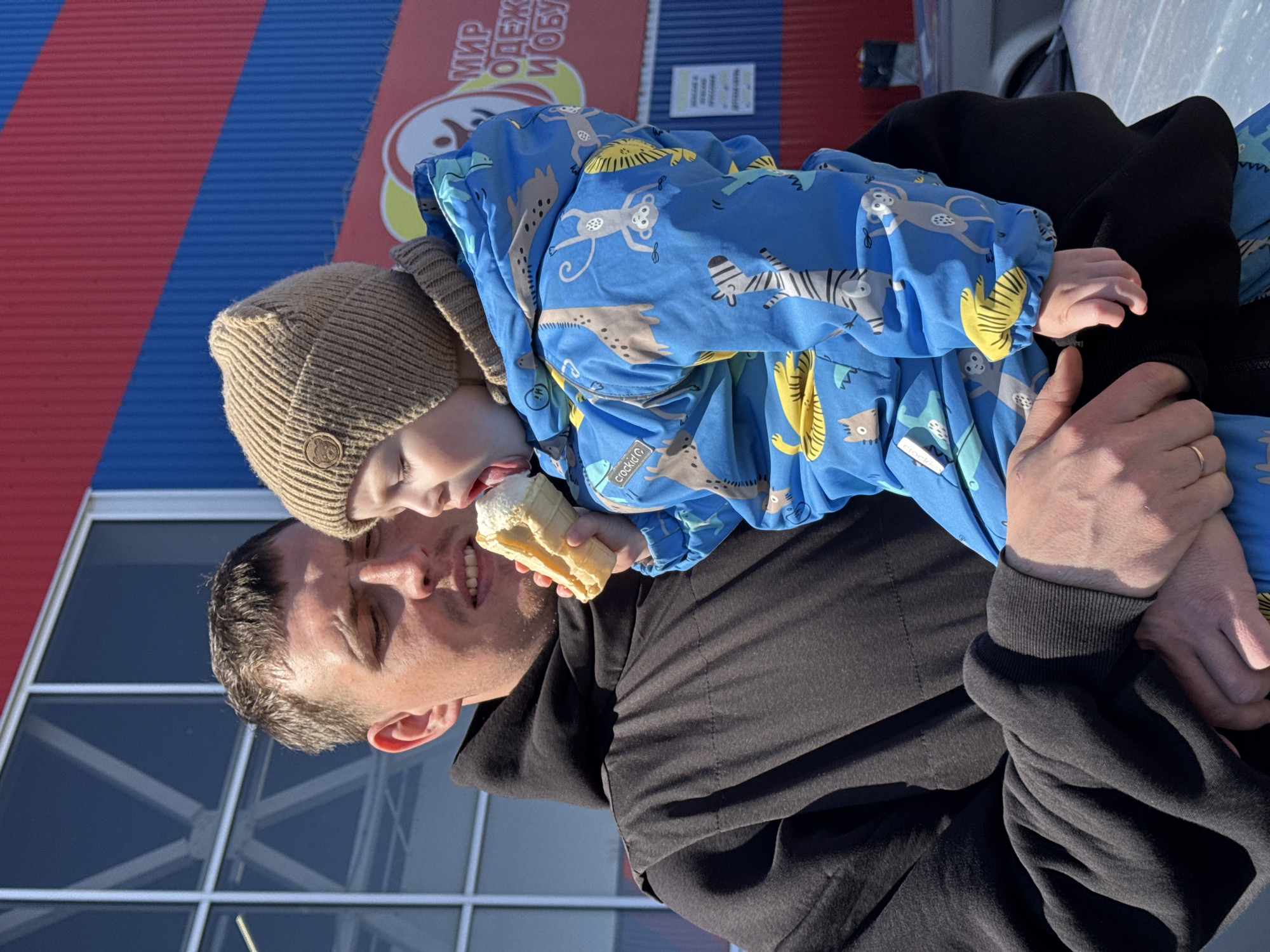 A light-skinned man and a small child stand outdoors in Chaikovsky, Russia. The man, wearing a black hooded sweatshirt, holds the child, who is dressed in a tan knit cap and a blue jacket with animal patterns. The child licks an ice cream cone. In the background is a building with red and blue stripes, a sign in Cyrillic, and windows. The scene is brightly lit by sunlight.