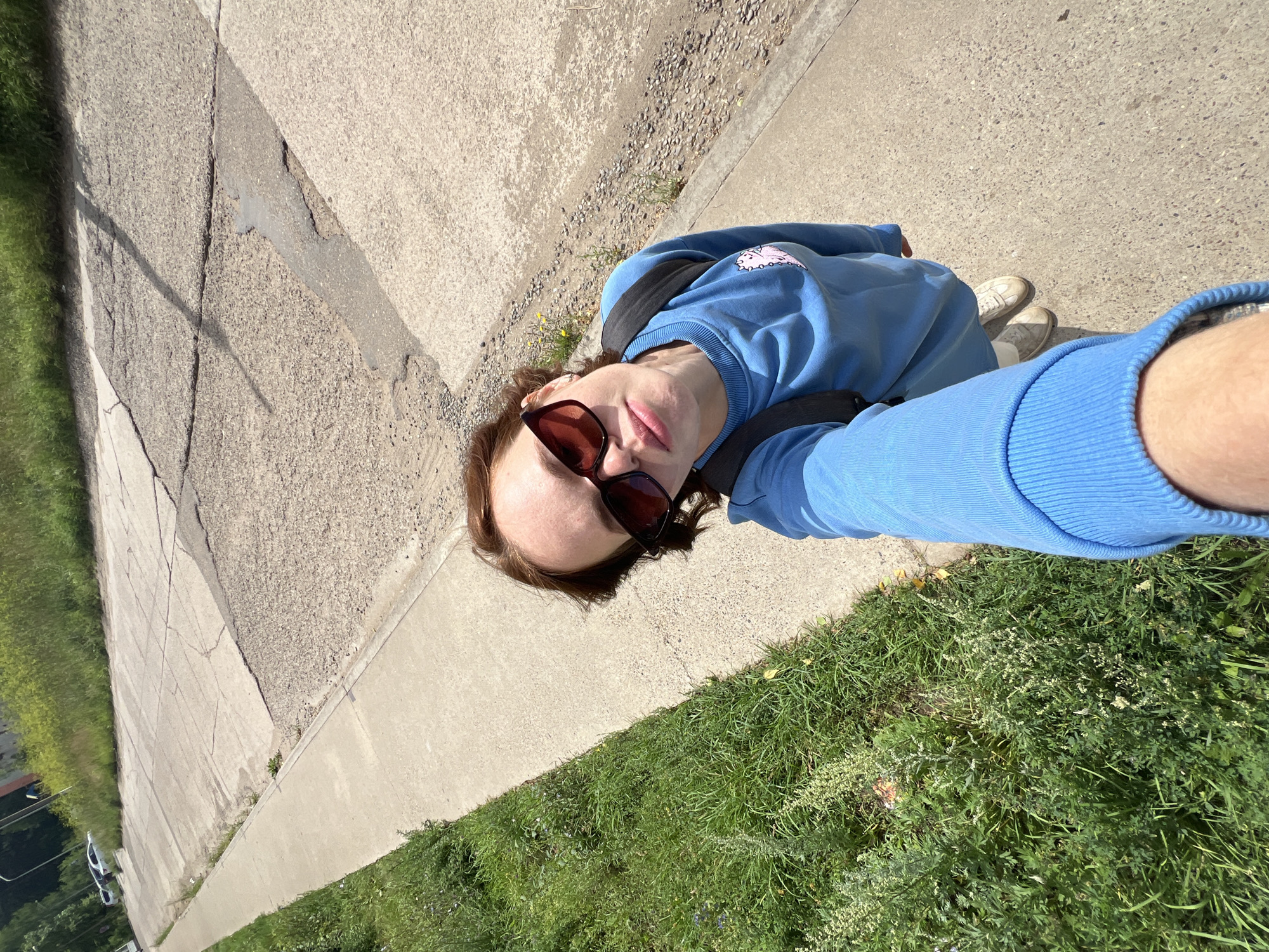 A young person, wearing sunglasses and a blue sweatshirt with a cartoon graphic, takes a selfie while standing on a paved sidewalk. They have a backpack on. The sidewalk runs alongside a grassy area and a cracked, multi-sectioned paved road. In the background, there is more grass, trees, and a few cars parked on the side of the road. The scene appears to be an urban or suburban environment.