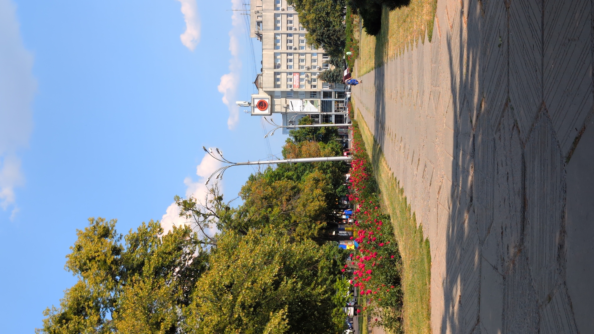 The image depicts a sunny day in Poltava, Ukraine, likely during daylight hours. The scene is an urban park or public walkway lined with trees and flowerbeds. A paved walkway, laid with patterned concrete tiles, dominates the right side of the frame, casting long shadows from unseen trees. To the left, lush green trees and a vibrant red flowerbed suggest a well-maintained public space.
In the background, a multi-story building with numerous windows stands tall, suggesting an urban setting. A prominent street lamp with multiple arms and lights stands near the center of the image, a common fixture in public areas. A sign with a red circle and a diagonal line across a number "4" is visible on a pole, possibly indicating a traffic regulation or informational notice. A person in blue is walking away from the camera on the path, while other people and parked cars are visible further down the street. The bright blue sky with scattered white clouds indicates fair weather. The overall impression is one of a pleasant, active outdoor space in a city.