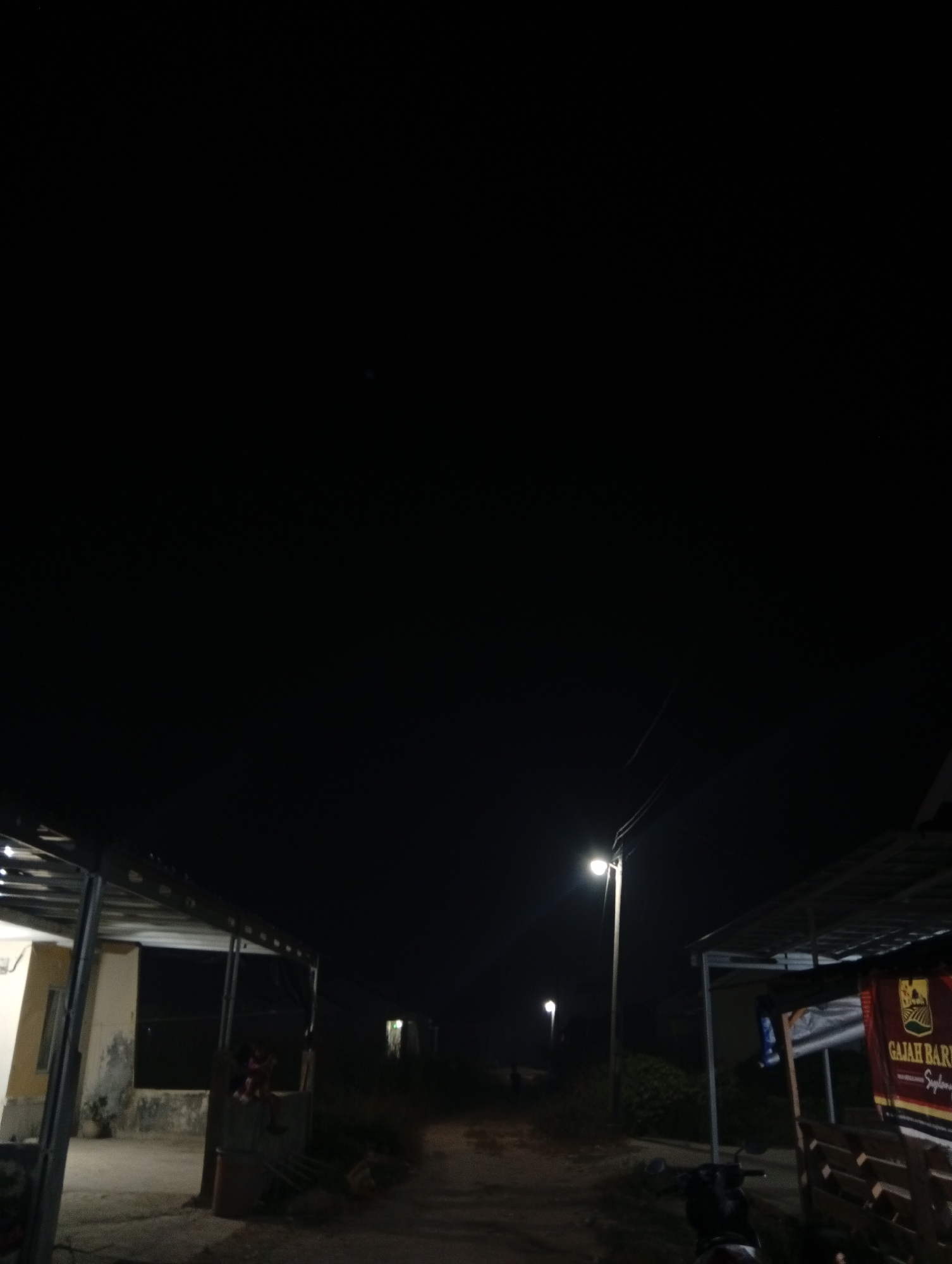 The image depicts an outdoor scene at night, likely in a rural or semi-rural area. The primary light source is a street lamp illuminating a dirt path. To the left, a building with a metal-framed roof structure and concrete walls is visible. A person is seated on a ledge in front of this building. To the right, another structure with a similar metal roof is present, and a banner with Indonesian text, "GAJAH BARU", is attached to it. A motorcycle is parked in front of this right-side structure. In the background, additional streetlights and dimly lit buildings suggest a small settlement or village. The overall impression is of a quiet, dimly lit street scene in Kualu, Indonesia.