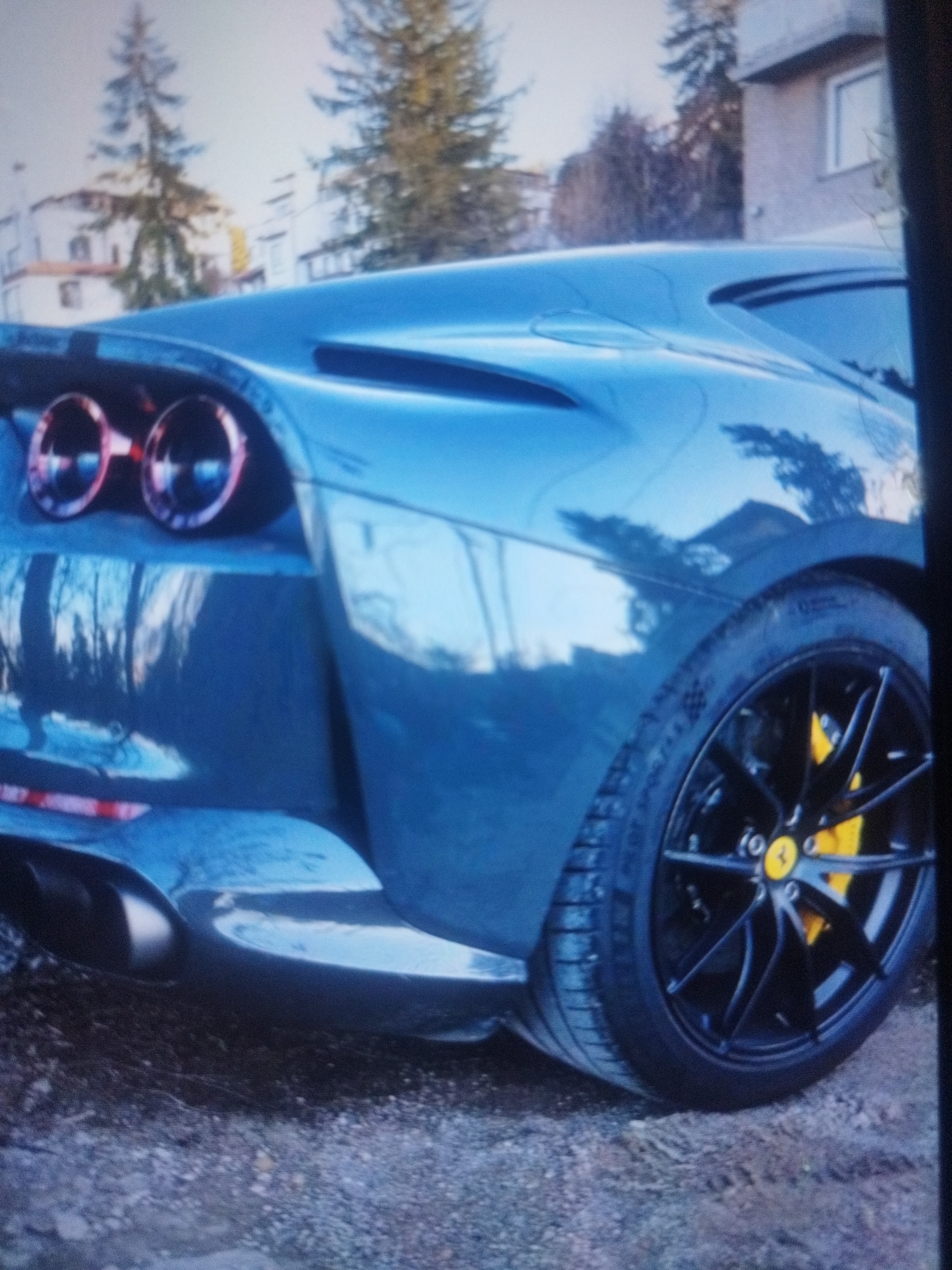The image shows the rear quarter of a dark blue Ferrari sports car parked on a gravel surface. The car features a black wheel with yellow brake calipers displaying the Ferrari logo. The taillights are round and have a red-orange hue. A double exhaust pipe is visible. The car's bodywork reflects the surrounding environment. In the background, buildings and trees are visible, potentially indicating a residential area. Given the prompt, the location is likely within the city of Plovdiv, Bulgaria, but this cannot be definitively confirmed from the visual evidence alone. No people are visible.