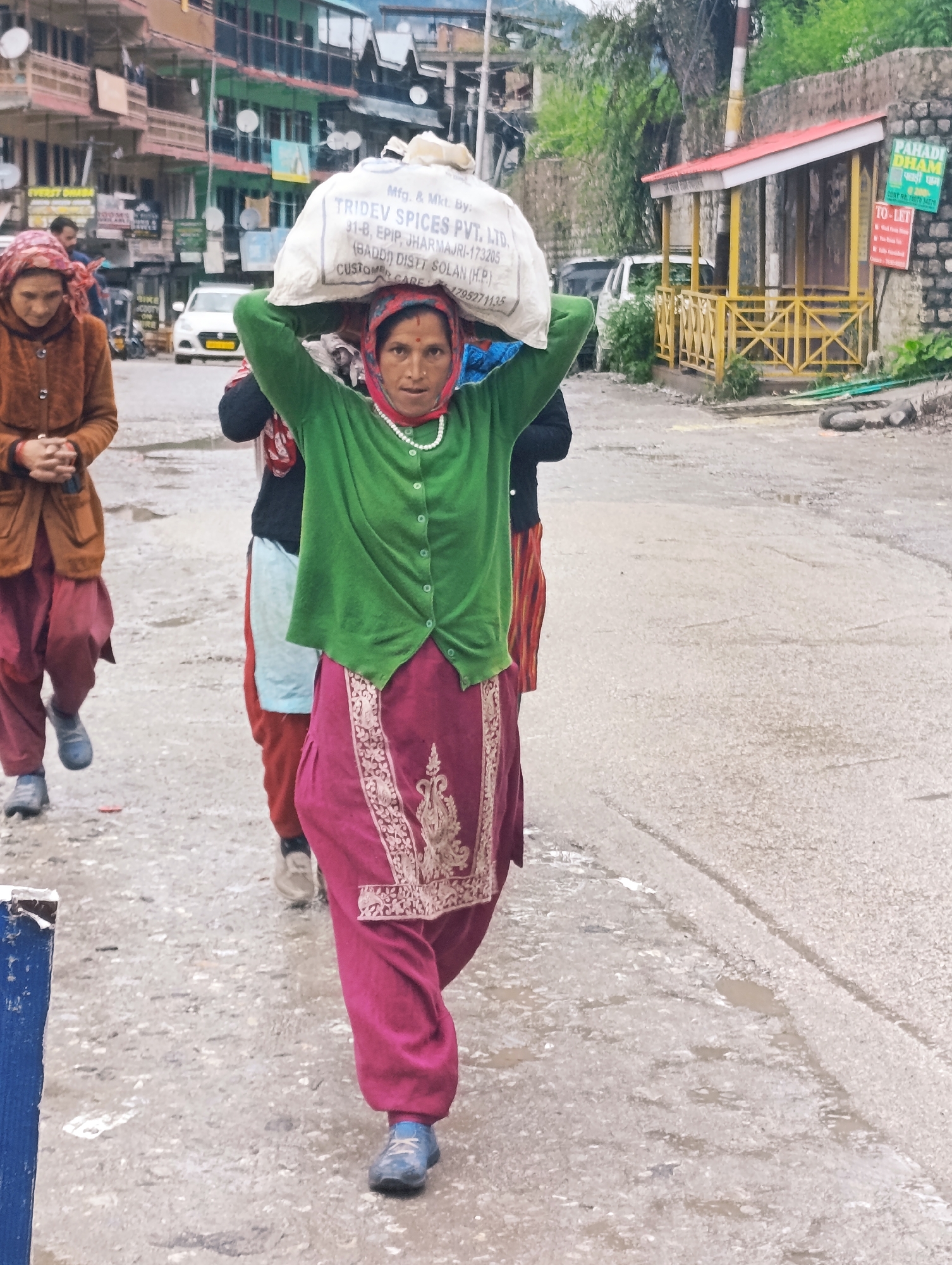 In Chichoga, India, three women walk along a wet, gravelly road. The woman in the foreground, wearing a green sweater and pink pants, carries a large white sack of "TRIDEV SPICES PVT. LTD." on her head. She is also adorned with a pearl necklace and a red headscarf. To her left, another woman in a brown jacket and pink pants walks with her hands clasped in front of her. A third woman, partially visible, walks behind the first woman. In the background, buildings with balconies line the street, and a white car is parked. A yellow structure with a red roof stands to the right, a sign reading "PAHADI DHAM" visible.