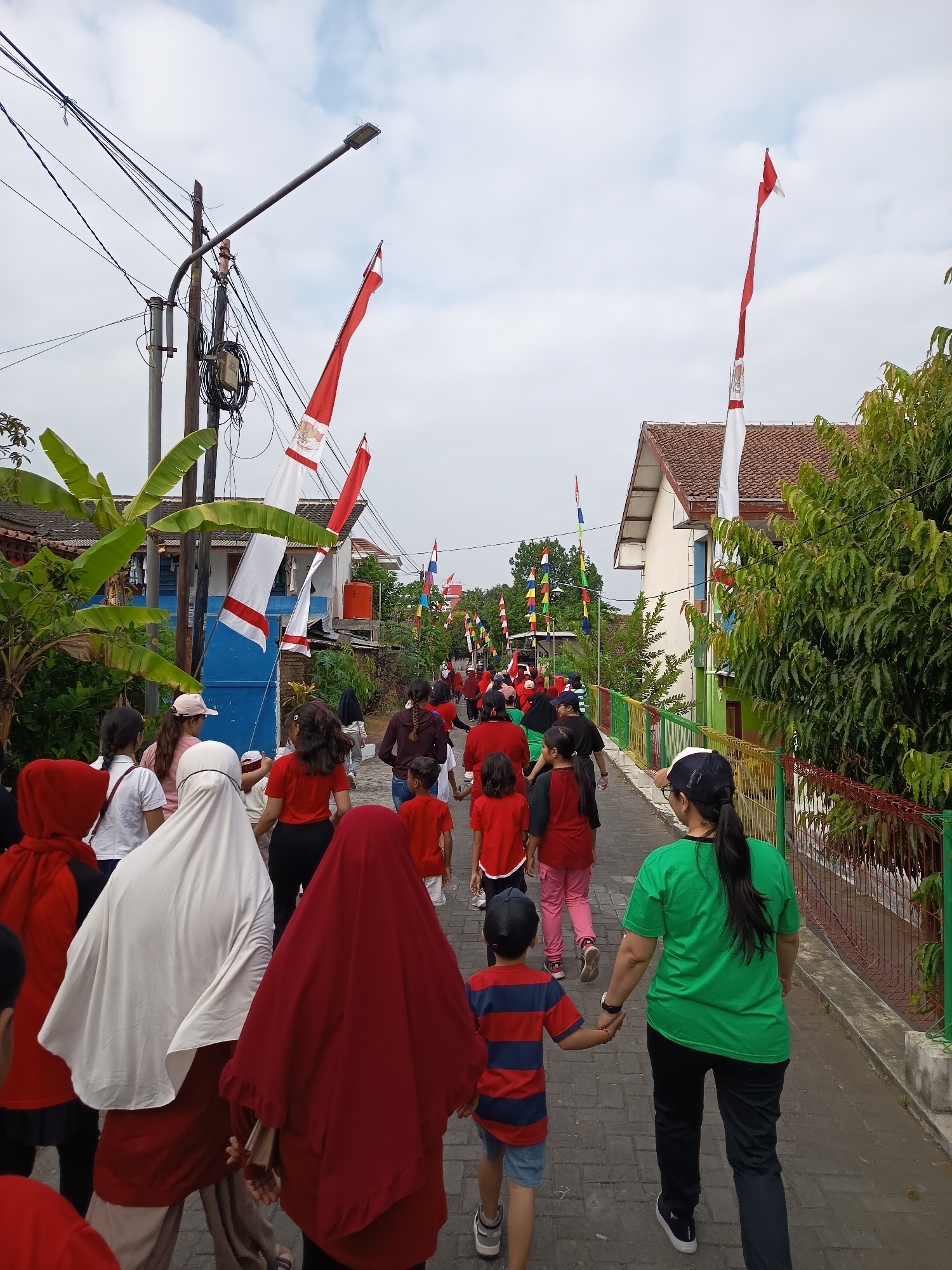 In the city of Ngaglik, Indonesia, a group of people, including men, women, and children, are walking down a paved street lined with houses and trees. Many of the people are wearing red and white clothing, and red and white flags are displayed along the street. The flags appear to be Indonesian flags. The sky is overcast. The street is made of brick pavers. There is a utility pole with wires in the foreground.