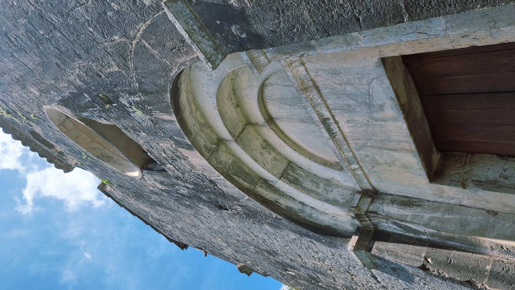 This is a low-angle, close-up, exterior shot of a stone structure in Sales de Llierca, Spain. The stone is dark gray and appears weathered, with some areas showing lighter green moss or lichen. The image is rotated so that the sky, which is a vibrant blue with scattered white clouds, occupies the lower portion of the frame.
The structure features an arched entryway, detailed with concentric curved stone moldings. To the left of this arch, a smaller, oval-shaped opening is visible in the stone wall, possibly a window or niche. The upper right portion of the image shows a section of a wooden door. The stone around the doorway is lighter in color and shows signs of age, with some cracking and discoloration.
The lighting suggests it is daytime, with bright sunlight illuminating the scene. The overall impression is of an ancient, possibly historical, building. The detailed stonework and the setting against the sky hint at an architectural feature of a church, castle, or similar edifice. There is no visible text in the image.
