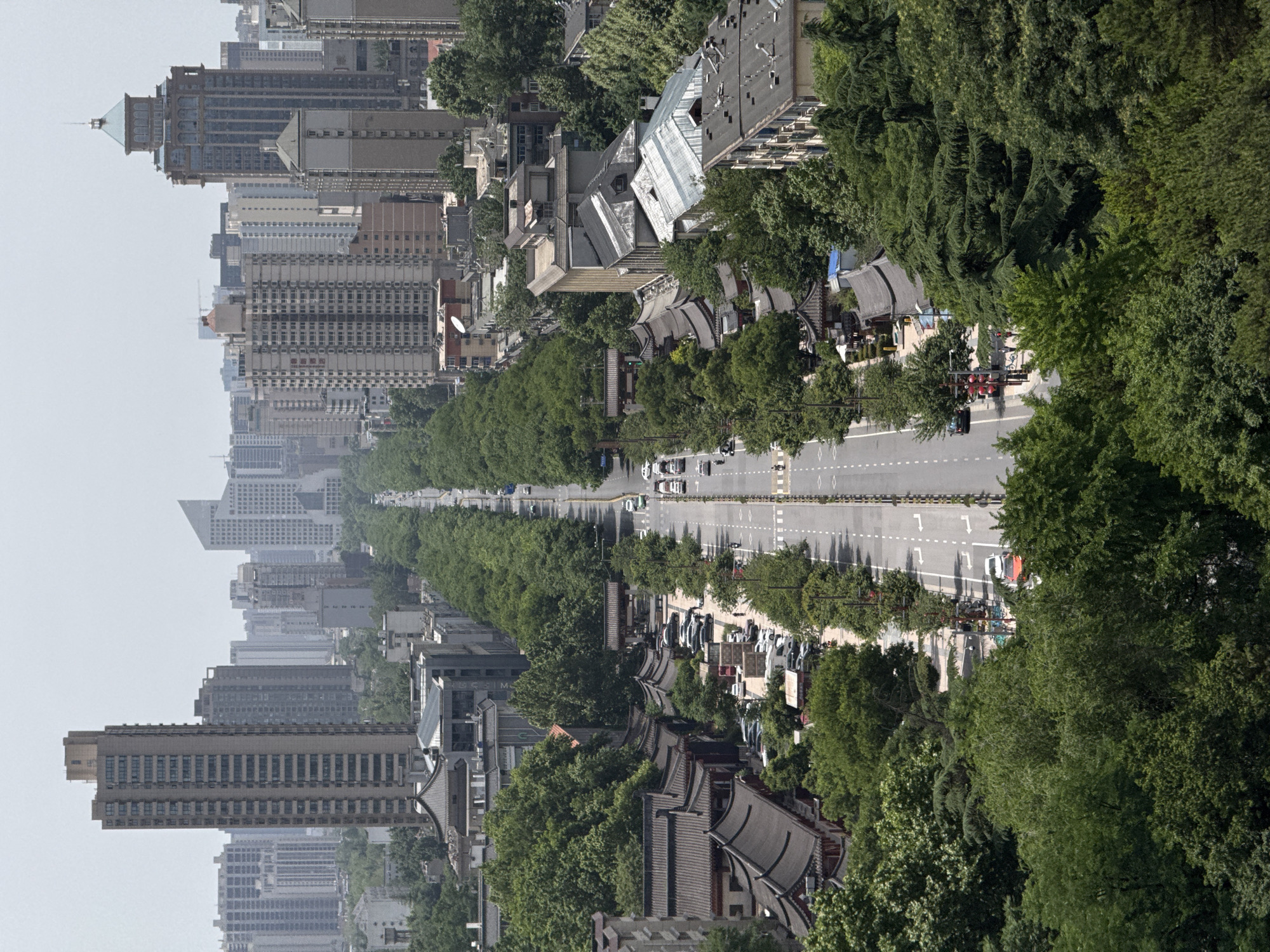 The image captures a street view in Yanta District, China, characterized by a blend of modern urban and traditional Chinese architectural elements. A straight, wide road lined with trees stretches into the distance, with cars moving along it. On either side of the road are buildings, with a mix of contemporary high-rise apartment buildings and traditional Chinese-style structures with curved roofs and ornate detailing. Trees and greenery fill the spaces between buildings, providing a natural contrast to the urban landscape. People are present, appearing as small figures near the traditional buildings, suggesting commercial or recreational activity in that area. The overall scene is set under a bright, slightly hazy sky, giving a sense of a clear day in a bustling urban environment with cultural preservation efforts.