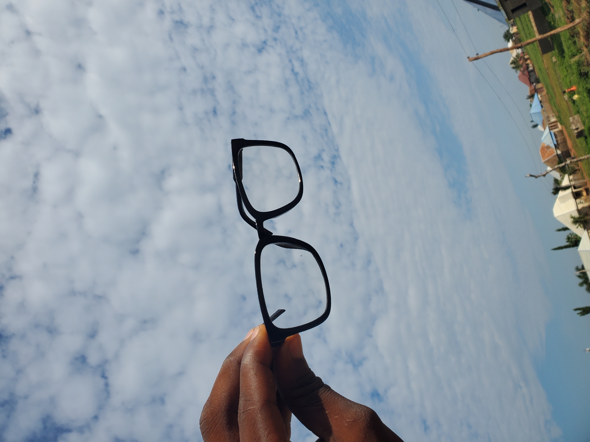 The scene is situated in the city of Winters, United States of America. A dark-skinned hand is prominently displayed in the upper left portion of the frame, holding a pair of black, rectangular-framed prescription glasses. The glasses feature clear lenses, with a small dark speck visible on the lens located on the viewer's right side. The background is dominated by a bright, light blue sky, extensively covered with numerous scattered white cumulus clouds, filling approximately two-thirds of the image vertically. The lower third of the image reveals a semi-urban or rural landscape under clear daylight. Several low-rise residential buildings with roofs of various colors, including blue, brown, and white, are visible in the mid-ground. Patches of green vegetation, likely grass, are interspersed among the structures and in the foreground. A wooden utility pole stands on the far right, and additional distant trees or poles are discernible. The image is composed from a low angle, looking upwards, causing the hand and glasses to be backlit against the bright sky.