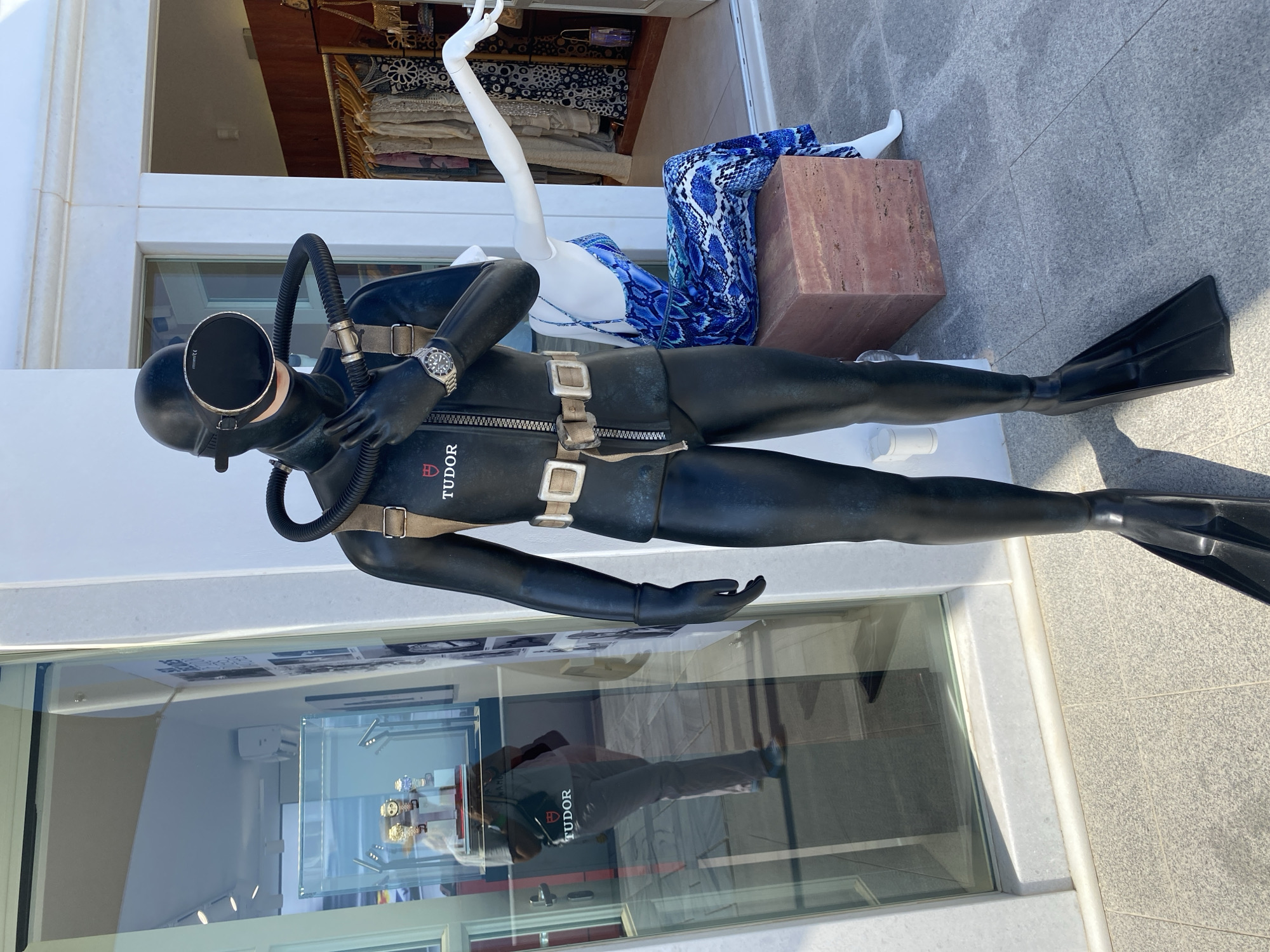 The image shows a life-sized mannequin dressed in a vintage black wetsuit with a diving mask and snorkel. The mannequin is positioned outside a storefront. A watch is visible on the mannequin's left wrist, and the word "TUDOR" is embroidered on the wetsuit. To the right of the mannequin, another mannequin is seated, wearing a blue and white patterned dress. Behind the seated mannequin, racks of clothing are displayed inside what appears to be a boutique. The storefront window on the left reflects a person inside the store, examining watches in a display case. The scene is set on a paved outdoor area, likely a sidewalk or plaza, with the background suggesting a sunny day in a coastal or urban environment. The building facade is white with architectural detailing.