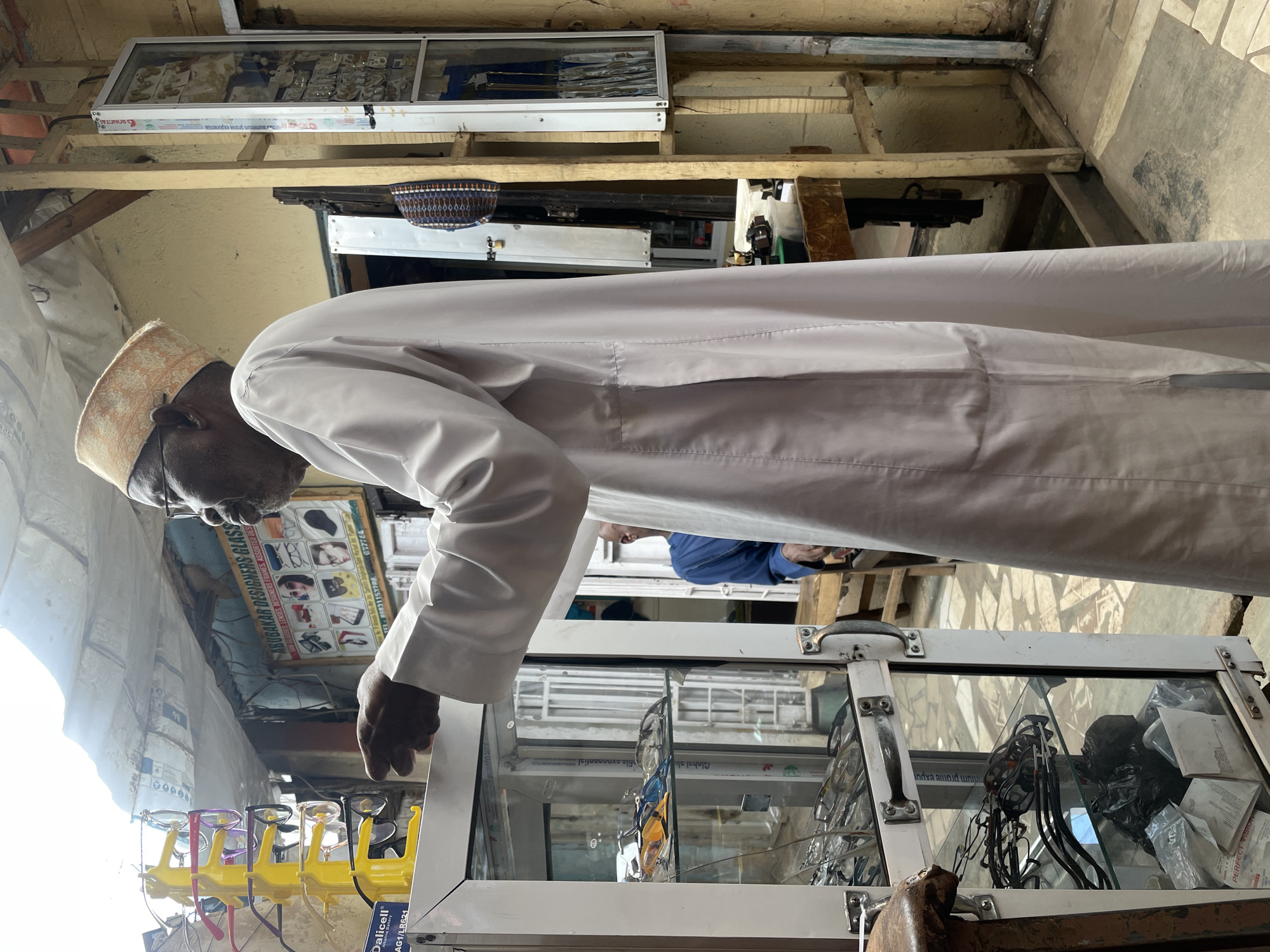 An elderly man with dark skin and wearing a light-colored, traditional patterned cap and glasses is looking at a display of eyeglasses in Jos, Nigeria. The glasses are displayed on bright yellow stands outside a shop called "Abubakar Designers Glass." The shop's sign also includes the phone number 637764 and the words "GLASSES LENSES DESIGNERS FRAMES ASSORTER DES." The man is wearing a long, light-colored robe. The shop is open-air and has a wooden frame. A display case with various items, including what appears to be lenses and other optical equipment, is located beneath the glasses display. Another man wearing a blue shirt is visible inside the shop.