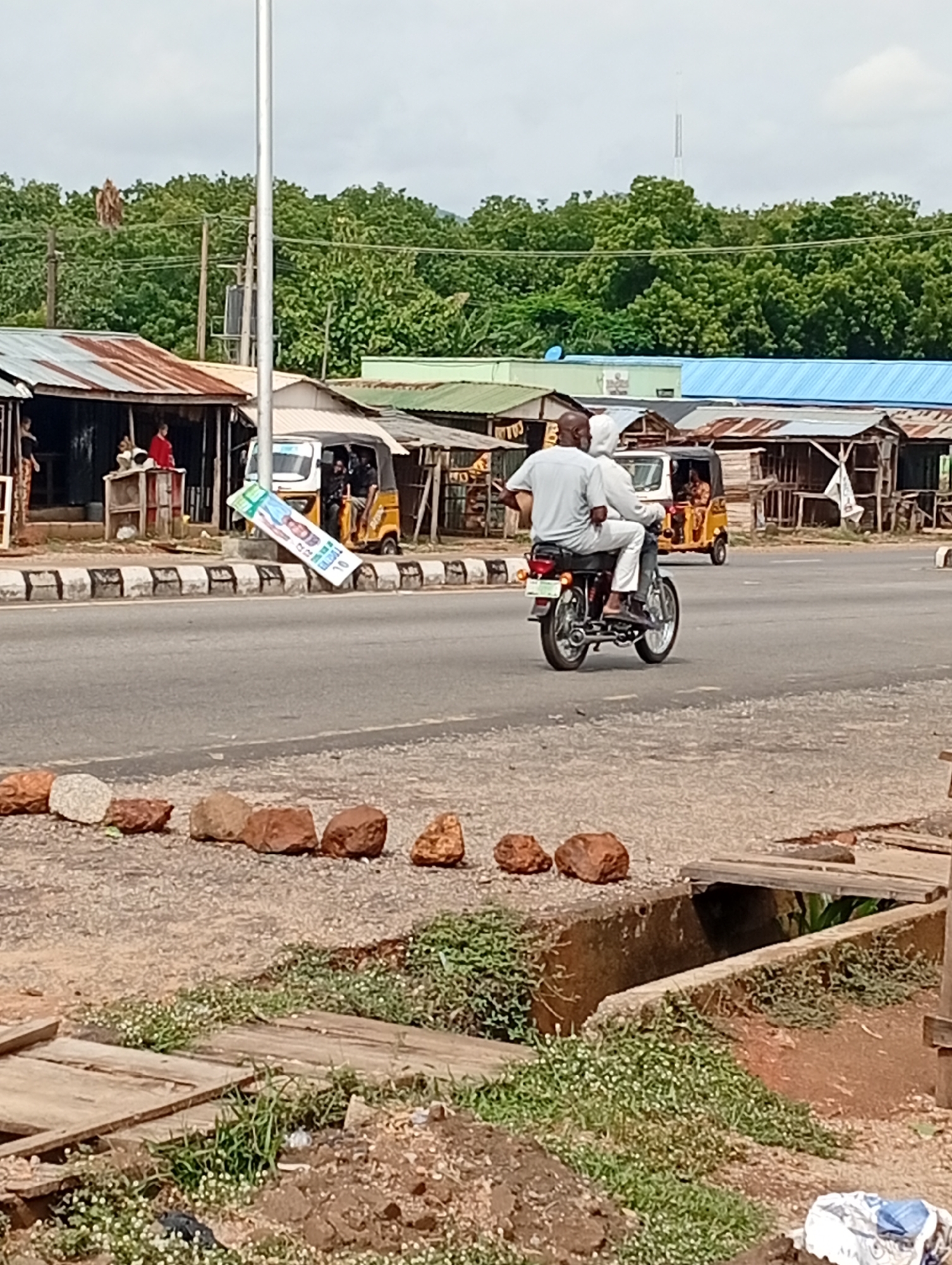The image depicts a street scene in Adankolo, Nigeria. Two people are riding a motorcycle on a paved road. The rider is wearing a light-colored shirt and dark pants. The passenger is wearing a light-colored outfit that includes a head covering. The motorcycle is moving from left to right in the frame.
Along the roadside are several small, open-front structures with corrugated metal roofs. Two three-wheeled vehicles (tuk-tuks) are visible near these structures. A vertical utility pole is also present near the structures. A political advertisement is seen on the roadside.
In the foreground is a dirt area with scattered rocks and low-lying vegetation. A concrete drainage ditch is covered with wooden planks, with additional vegetation growing nearby. Trees and other greenery are visible in the background.