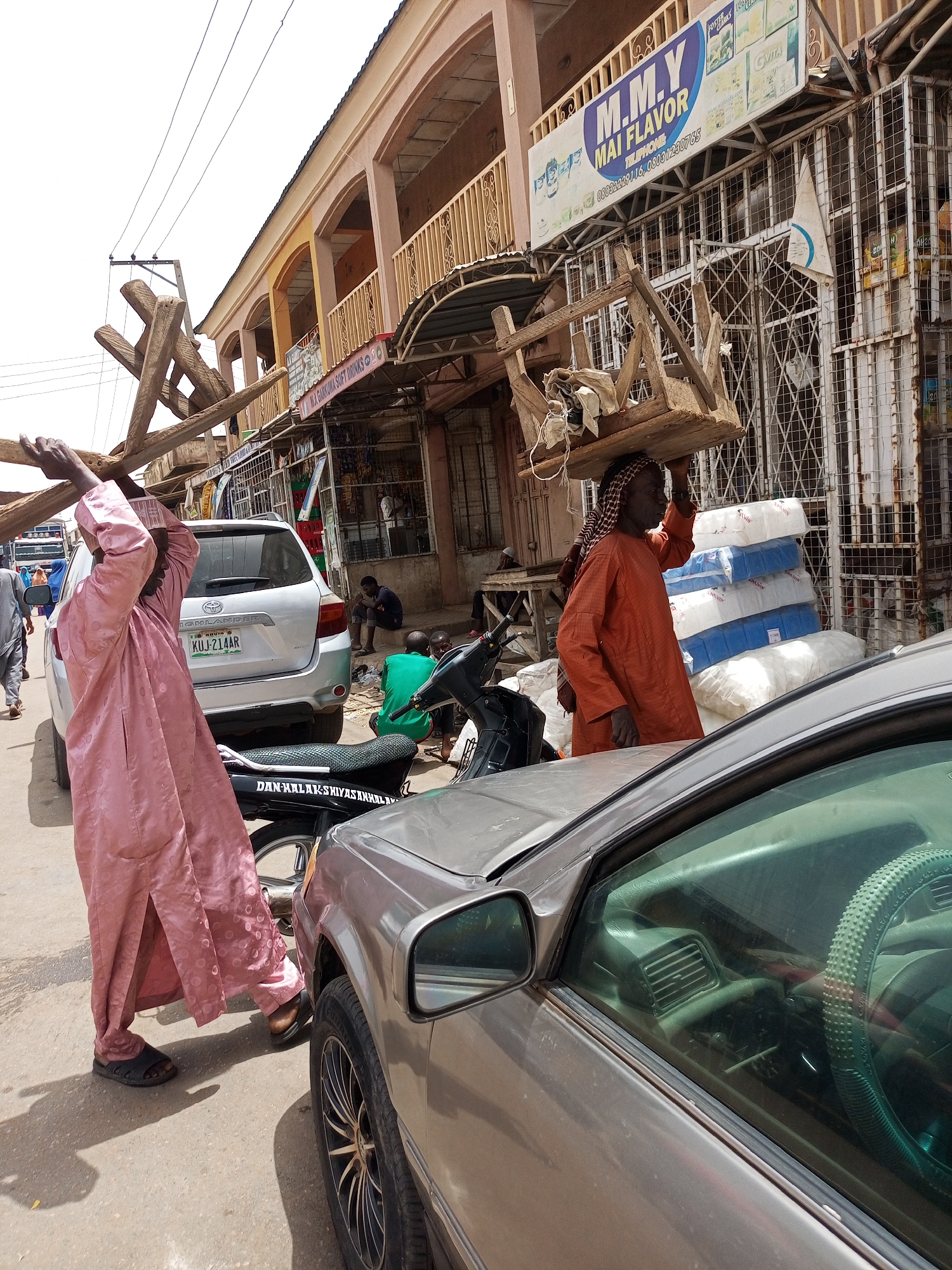 The scene is a street in Arawa, Nigeria, in front of shops. A man in a pink robe and hat carries wooden planks balanced above his head. A second man in an orange robe and patterned head covering balances a wooden object on his head, and is standing near stacks of packages. The vehicles are a silver Toyota Highlander with license plate "KUJ-214AR", a black motorcycle, and a gold car. A building with a sign for "M.M.Y MAI FLAVOR" stands behind the men, alongside other shops. Several individuals are seated on the sidewalk. Overhead electrical wires crisscross the sky. The scene has a street-market atmosphere.