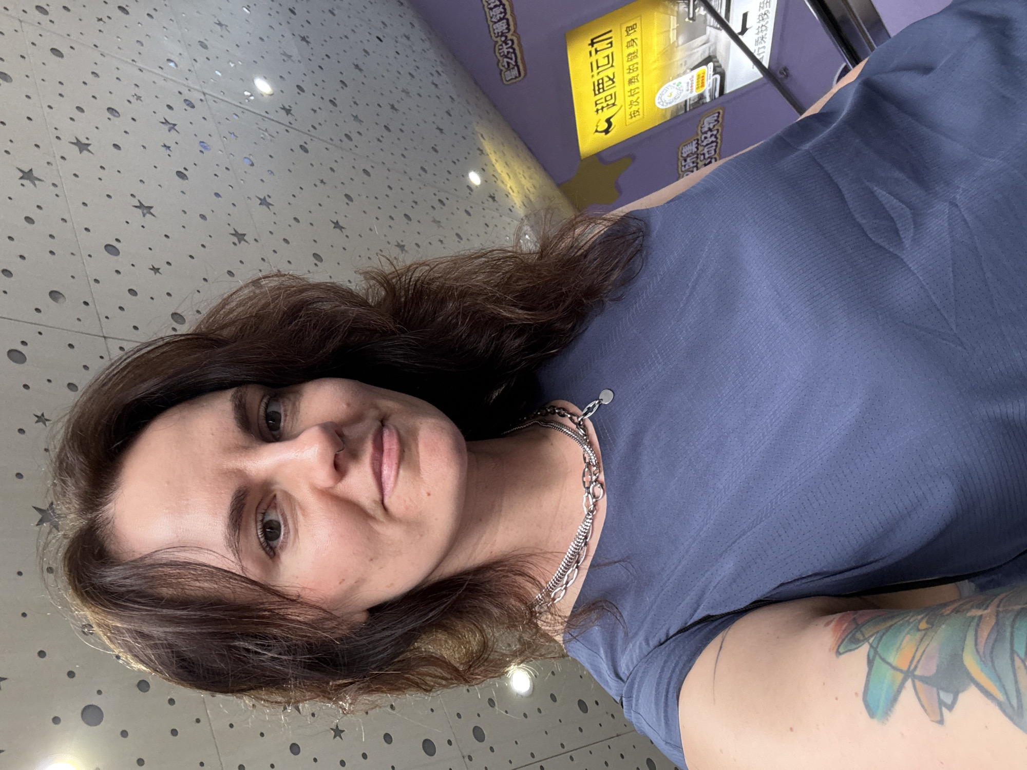 The image shows a woman with brown hair and a blue shirt taking a selfie. She has a silver chain necklace and a tattoo on her upper arm. In the background, there's a sign in Chinese characters with the text "超鹿运动 (Chao Lu Yun Dong)" and "按次付费的健身馆 (An Ci Fu Fei De Jian Shen Guan)", which translates to "Super Deer Sports" and "Pay-per-visit Gym". The first line on the same sign is 星之光・潮领址, which indicates a location. The sign suggests she might be in a gym or a fitness center. The wall behind her has a pattern of dots and stars. Given that the sign is in Chinese, it is likely she is in a Chinese-speaking region. The context suggests that the location may be a fitness center in the Tianhe District of China.