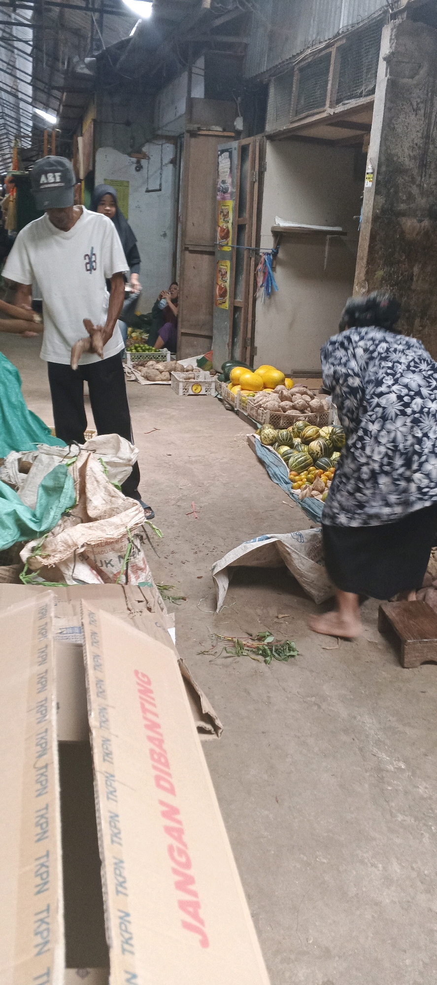 The image depicts an outdoor market scene, likely in Sekaran, Indonesia, with a focus on the sale of produce. In the foreground, a man wearing a white t-shirt, black pants, and a grey baseball cap is holding a piece of wood. To his right, a woman in a dark floral-patterned dress and black skirt is bending over, possibly examining produce. Various fruits and vegetables are displayed on the ground and in crates, including what appear to be squashes, watermelons, yellow melons, and root vegetables. Cardboard boxes are stacked in the lower left portion of the image, one of which has text visible that includes "JANGAN DIBANTING". In the background, other individuals can be seen, and the market stalls are constructed from wood and corrugated metal, suggesting a rustic and possibly covered environment. The ground is concrete or dirt.