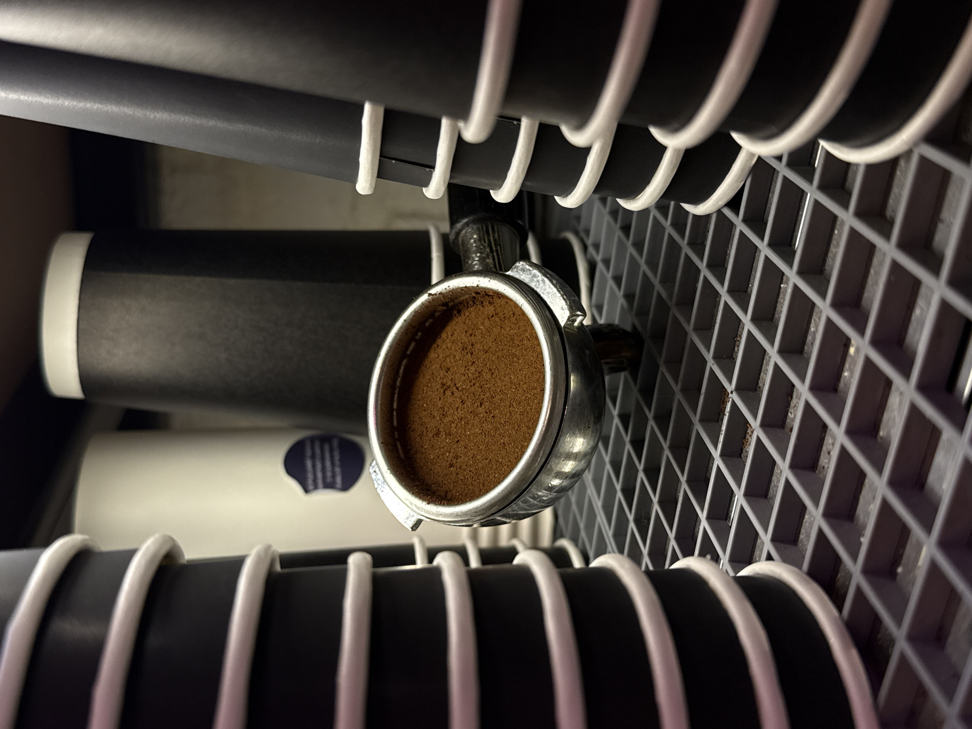 The image shows a close-up view of a portafilter filled with finely ground coffee, placed on a grey plastic grate. In the background, several stacked black paper cups with white rims are visible. To the left, another stack of white paper cups with black rims is also present. The lighting appears to be artificial, casting shadows and highlighting the texture of the coffee grounds. The scene is clean and organized, suggesting a professional coffee preparation environment. The specific location in the city of Dolgoprudny, Russia is not discernible from the image itself.