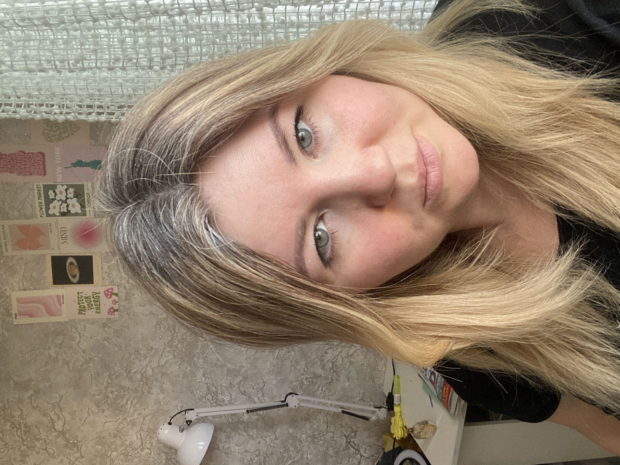A close-up, slightly tilted portrait shows a person with long, blonde and graying hair, looking directly at the camera with light green or hazel eyes. The subject has fair skin and light pink lips. The photo appears to be a selfie taken indoors.
In the background, a light-colored, textured woven fabric or curtain is visible at the top, likely behind the subject's head. Below this, a wall is adorned with several small, rectangular prints or postcards. To the lower left, a white articulated desk lamp is positioned on a surface. Further to the right, behind the subject's hair and shoulder, parts of a desk or shelf are visible with indistinct items.
Notable details include the striking contrast between the blonde and gray hair. The subject's expression is neutral and direct.
Visible text on the wall posters includes: "FLOWER MARKET", "NEW YORK", "LONDON", "MIND", "PROTECT YOUR ENERGY", and "PAPIERS DECOUPES". There is also Cyrillic text, including "ТИПОВЕ" (potentially part of "типовое" meaning 'typical') and "СЕ ПРЕ" (likely part of a larger word or phrase). The word "Fisk" is also seen. One poster features an image of Saturn, and another shows mushrooms. Partially visible Cyrillic text like "ОБОВ" on items on the desk hints at the given location context of Chaikovsky, Russia.