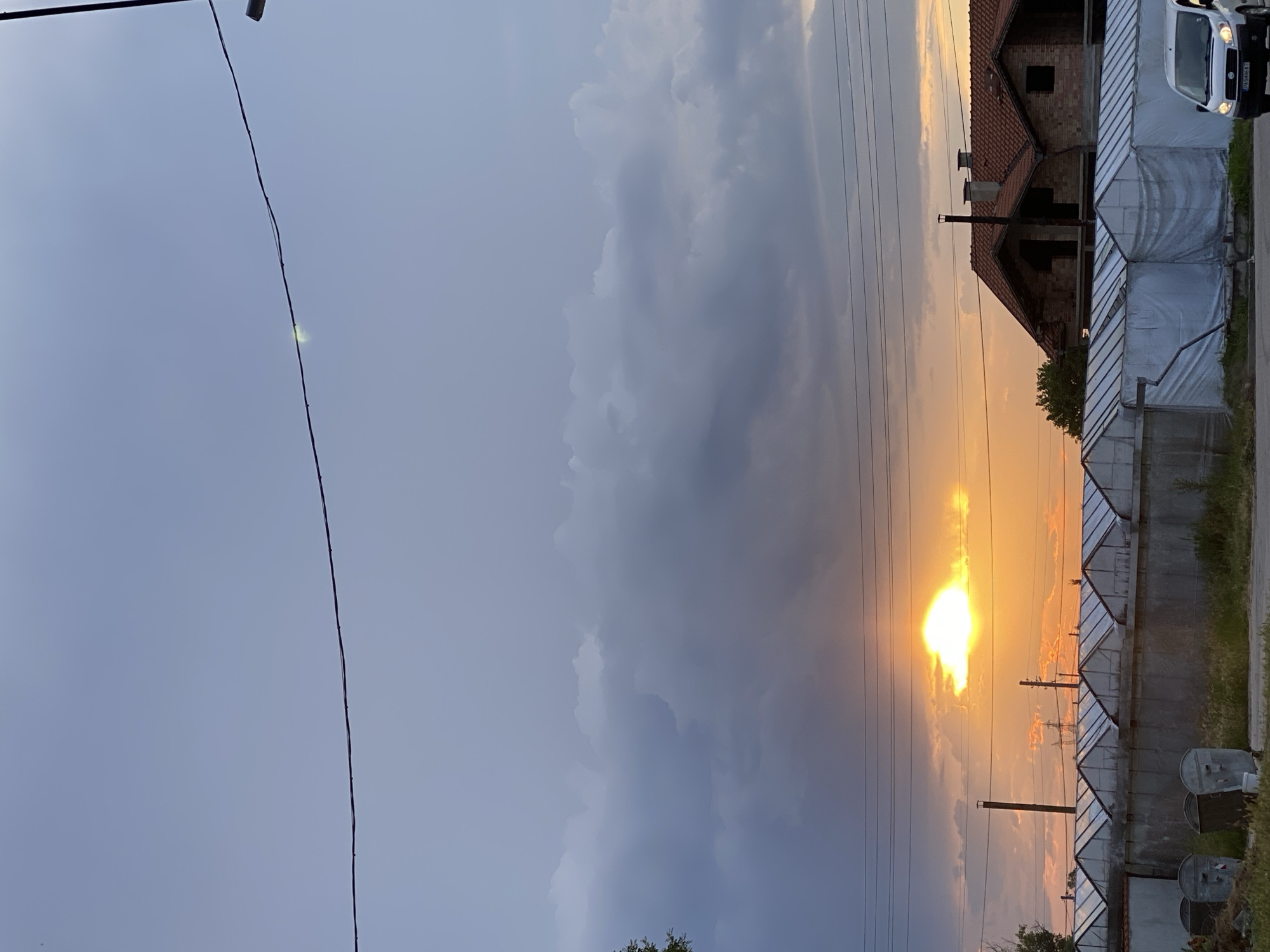 A vibrant sunset illuminates a rural or semi-rural scene in Miriantsi, Bulgaria, captured during dusk. The image is dominated by a dramatic sky, split between deep blue-gray on the left and intense orange and yellow hues on the right, where the sun glows brightly through and behind clouds. The sun itself is a powerful, diffuse light source, partially obscured by thick, dark clouds that reflect its warm colors.
Several dark electrical wires stretch horizontally across the upper and middle sections of the frame, contrasting against the colorful sky. A faint, small green-yellow light or reflection is visible on one of these wires towards the upper left. On the far right, a two-story brick house with a red tiled roof appears to be under construction or unfinished, featuring dark, empty window openings and a prominent chimney. Adjacent to and in front of the house are multiple structures resembling greenhouses or covered sheds, characterized by concrete block bases and translucent white, sloped coverings. These structures extend across the middle-right of the image, below the house. A utility pole is also visible amidst these structures.
A white van is partially visible parked on the far upper right, with a license plate displaying "B 544AX" (partially legible). In the lower right corner, two metallic garbage cans or large bins are situated near the edge of the property. Sparse vegetation, including trees and grass, can be seen near the buildings and along the ground. No human activity is depicted, focusing instead on the atmospheric landscape and the interplay of natural light with man-made elements.
