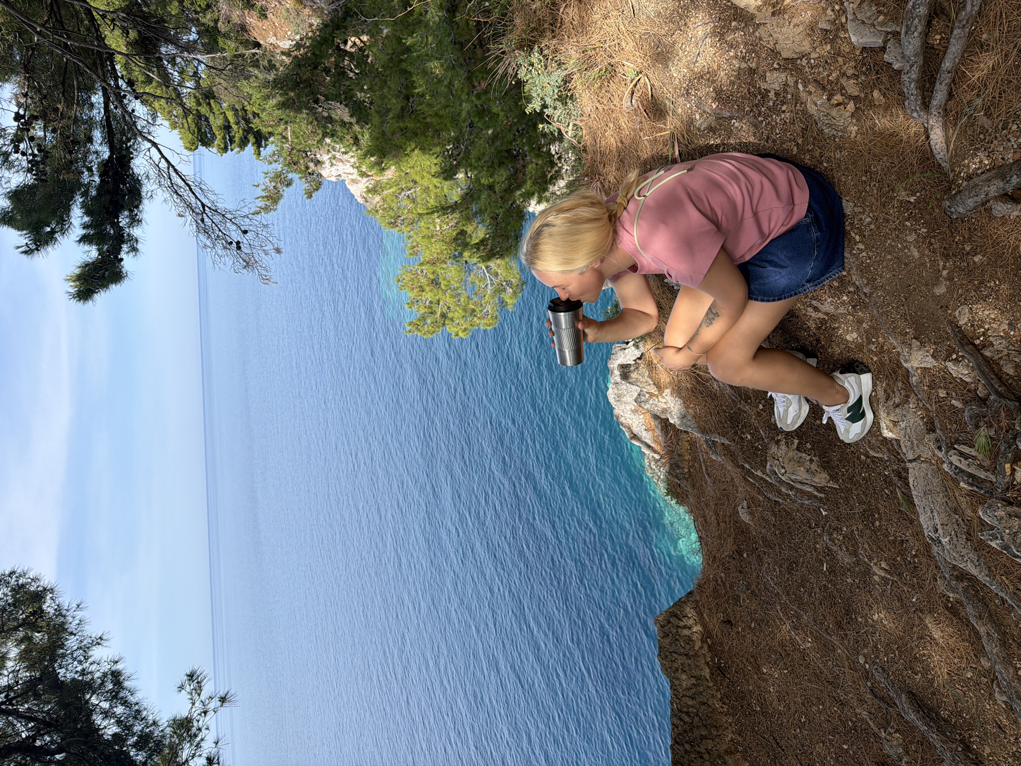 A woman with blonde hair in a pink t-shirt and blue shorts is kneeling near the edge of a cliff. She is drinking from a stainless steel mug. The cliff overlooks a body of water. Trees and foliage are visible on the cliffside. The photograph appears to have been taken in Petrovac na Moru, Montenegro, based on the location. The ground is covered in pine needles and roots. She is wearing white and green sneakers. A tattoo is visible on her left arm.