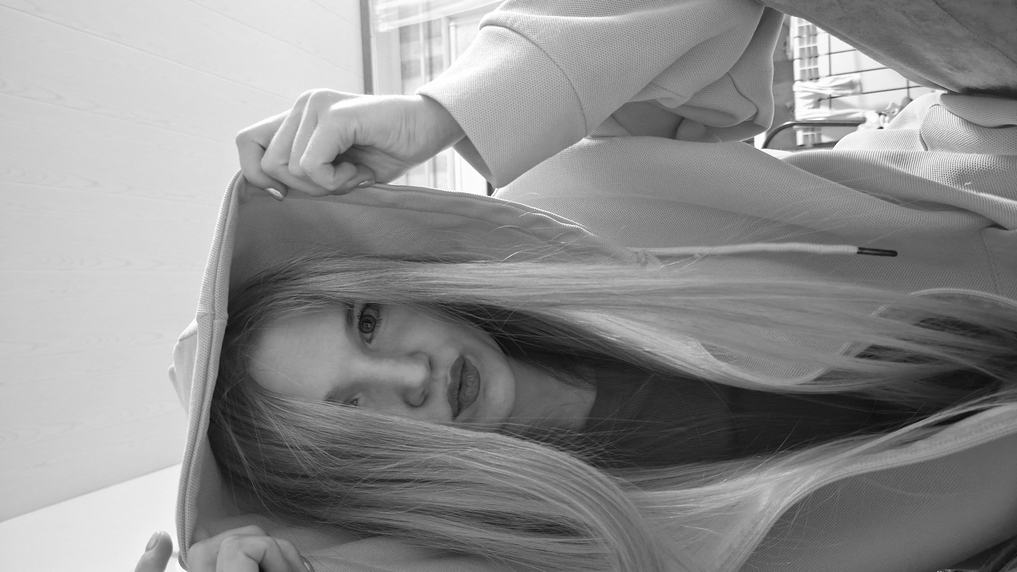 This black and white portrait features a young individual, likely female, with long, light-colored hair, looking directly at the viewer from a tilted perspective. The person is wearing a light-toned hoodie, with their hands visible holding the edges of the hood, partially obscuring the face. One eye, part of the nose, and the mouth with slightly visible lips are distinguishable through the curtain of hair and the hood. The visible eye displays a direct and engaging gaze.
The hoodie appears to be made of a textured, possibly knit, fabric, and a drawstring with a dark metal tip can be seen on the garment. The background consists primarily of a light-colored surface with horizontal paneling, suggesting an indoor wall or ceiling. In the upper right section of the frame, a blurred window or framed structure with a grid pattern is visible, indicating an indoor setting with natural light filtering in. The time of day appears to be daytime. No text is visible in the image. The overall impression is intimate and slightly mysterious due to the partial concealment of the face.