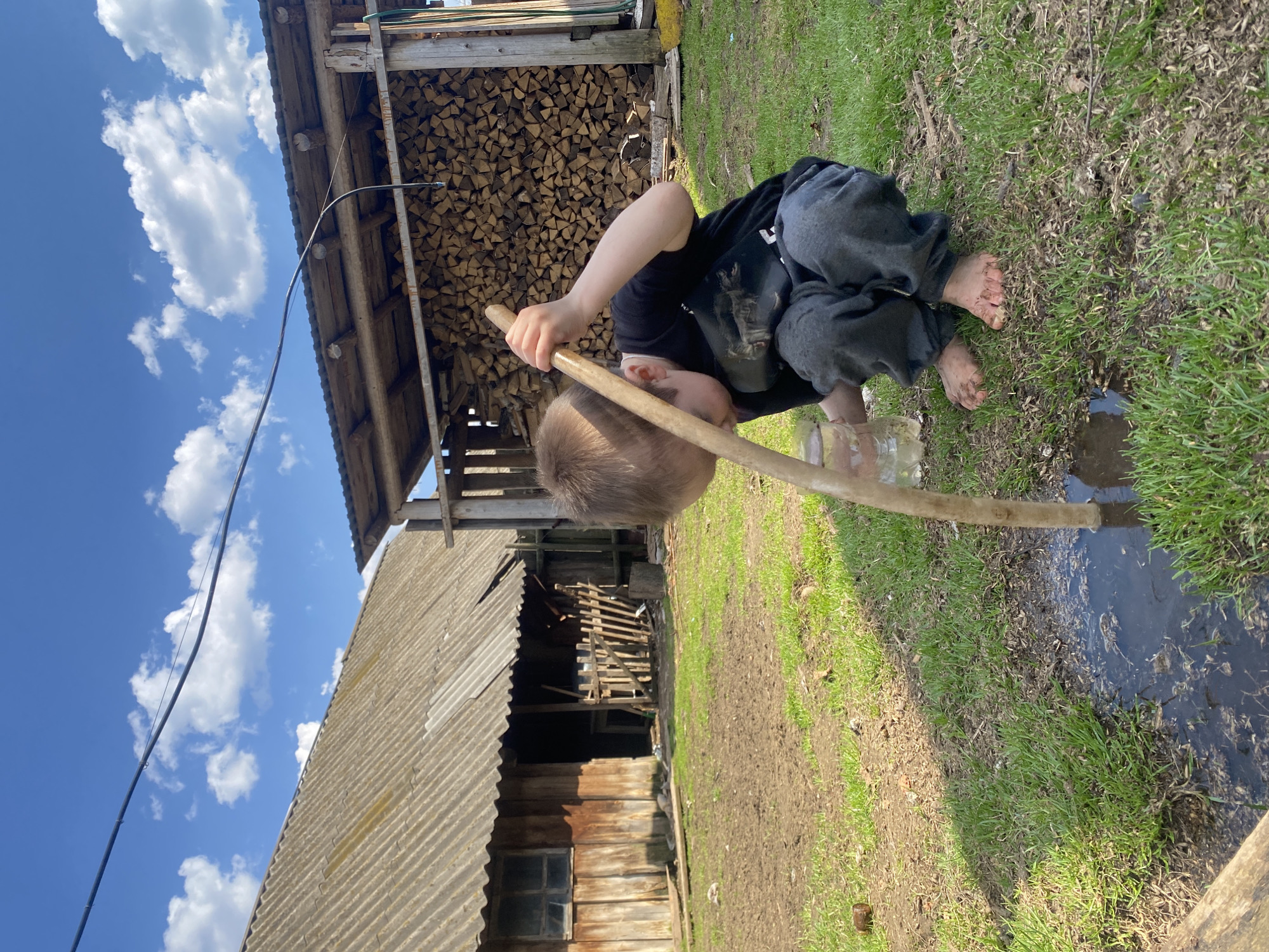 A barefoot child is squatting by a puddle, holding a stick in their hands. They are wearing a black t-shirt and grey sweatpants. Behind the child is a stack of firewood and an old wooden building with a corrugated metal roof. The sky is blue with white clouds. The ground is a mix of grass and dirt. The location is consistent with the rural characteristics of Узяр, Russia.