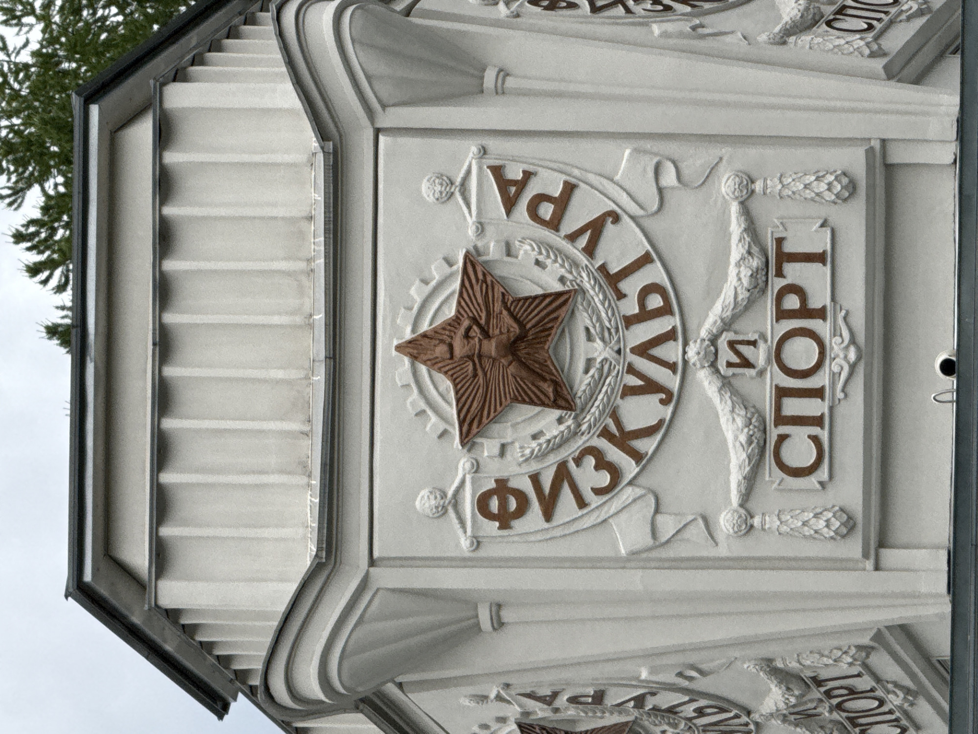 The image shows a close-up of a building facade, likely in Moscow, Russia, featuring relief sculptures and text. The main subject is a circular emblem with a five-pointed star at its center. The star is brown and has intricate detailing. The emblem is surrounded by text in Cyrillic letters. The upper half of the text reads "ФИЗКУЛЬТУРА" (Fizkultura), meaning "Physical Culture," and the lower half reads "И СПОРТ" (I Sport), meaning "and Sport." This suggests the building is related to sports or physical education.
The relief sculpture is made of a light-colored material, likely plaster or stone, and is set against a textured background of horizontal lines, possibly representing a facade or a decorative element of the building. Flanking the circular emblem are decorative elements resembling stylized torches or pinecones. The overall style of the relief suggests a Soviet-era architectural influence.
In the upper left portion of the image, a portion of a green conifer tree is visible against an overcast sky, indicating an outdoor setting. The lighting appears to be natural daylight, and the weather seems to be cloudy. The perspective is slightly tilted, giving a dynamic feel to the composition. No people are visible in the image.