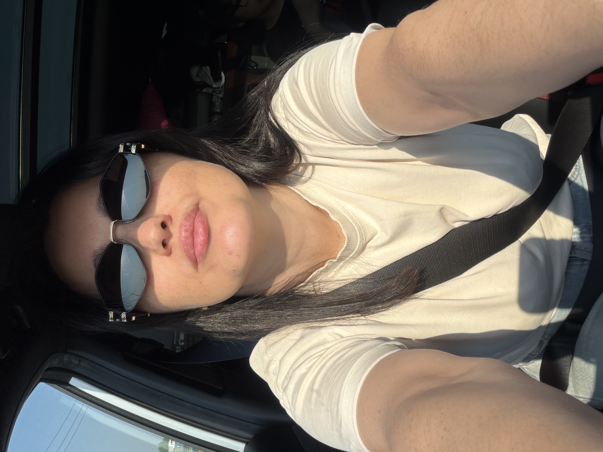 A woman, with long dark hair and sunglasses, takes a selfie while sitting in the driver's seat of a car. She is wearing a light-colored t-shirt and a seatbelt. The sun is shining brightly, casting shadows on her face and arm. The interior of the car is dark, with the sunroof open. A person is visible in the passenger seat in the background. The image does not provide enough information to determine the location within the city of Bolshiye Dvory, Russia.