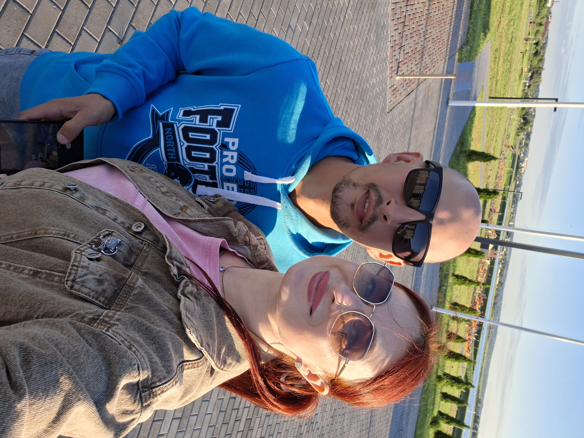 The image is a selfie taken outdoors in a city park in Smaliavichy, Belarus. In the foreground, a man and a woman are posing for the camera. The man is wearing a blue hooded sweatshirt with a "Pro Football North" logo and sunglasses. The woman has red hair pulled back in a ponytail, is wearing sunglasses, a pink t-shirt, and a denim jacket. The background shows a paved walkway, a grassy hill with trees and some playground equipment, and a body of water. The sky is clear and blue.