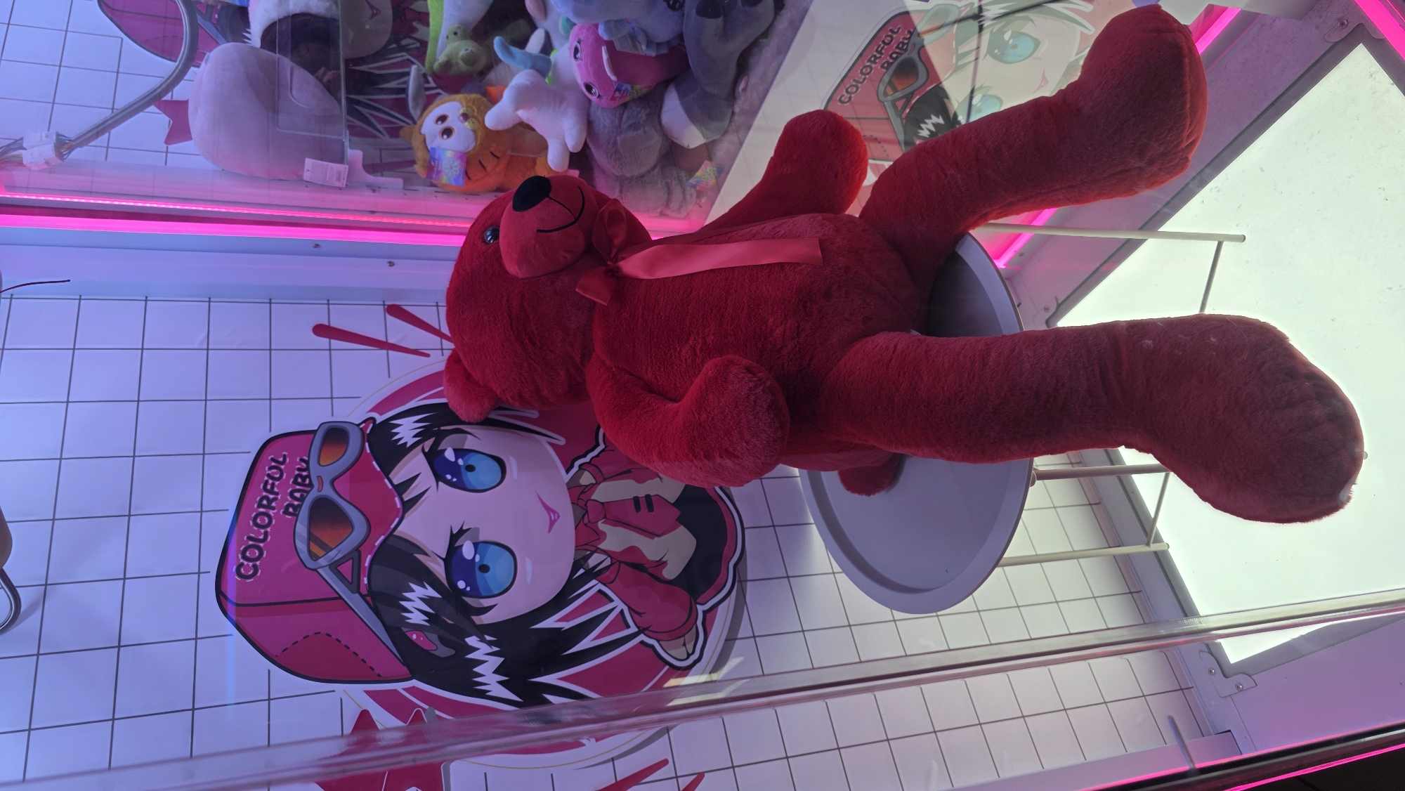 A red plush teddy bear lies atop an anime-style sticker inside a claw machine. The machine is in a brightly lit, grid-tiled environment. Other plush toys are visible in the background. The sticker features a character with large blue eyes and a pink hat labeled "COLORFUL BABY". The bear is positioned over a gray saucer, possibly a prize platform. The claw machine’s frame is visible in the foreground. There is no specific information to indicate that this image was taken in Blagoevgrad, Bulgaria.