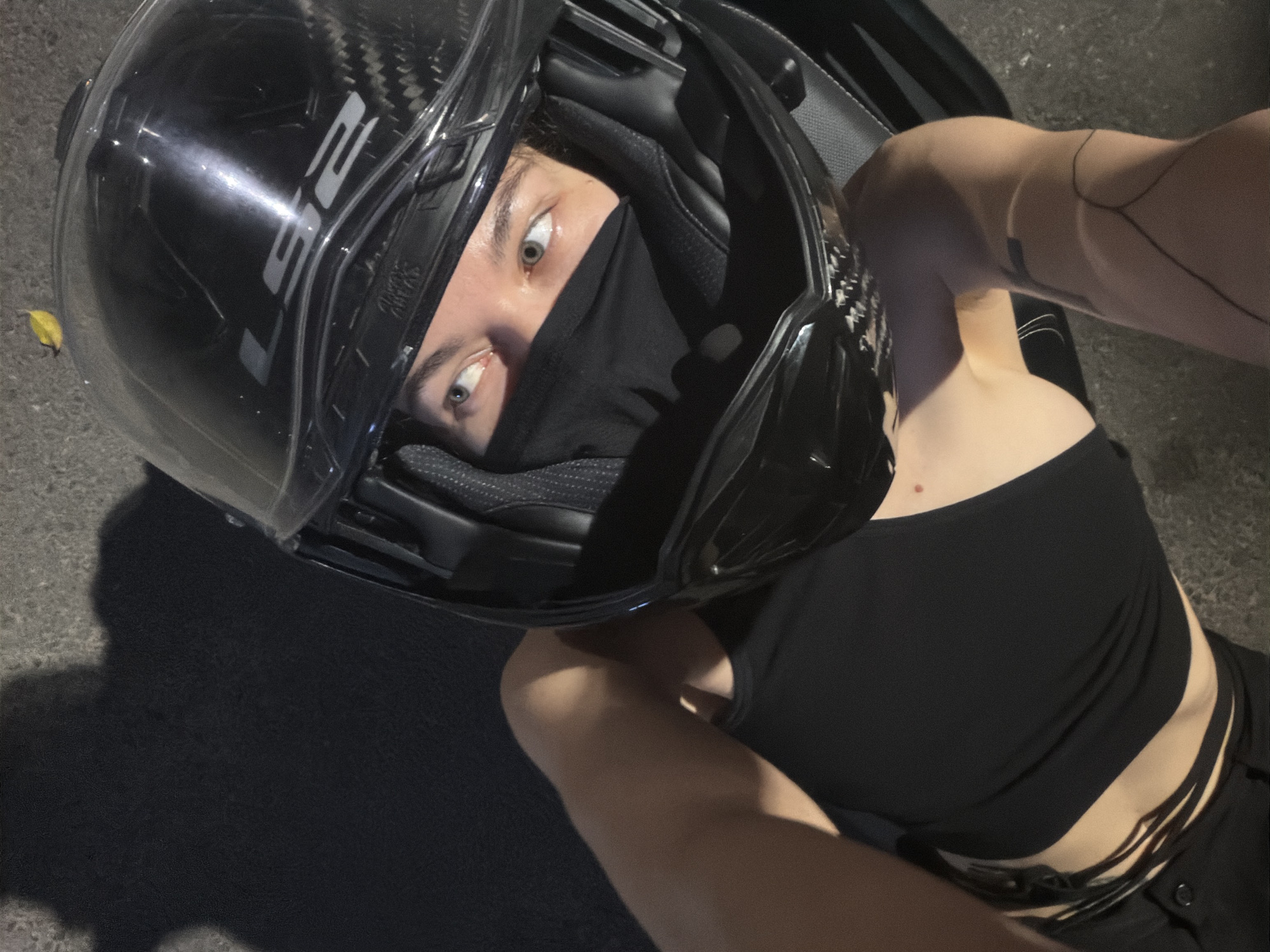 The image captures a person wearing a dark, possibly carbon-fiber patterned, full-face motorcycle helmet with the brand "LS2" displayed in silver/white lettering on the side. A black face mask covers the lower half of the person's face, leaving only their wide, light-colored eyes visible, which convey an intense or surprised expression. The individual is wearing a black, strapless-style top and dark bottoms with decorative straps around the waist. They appear to be lying or leaning on a dark, rough-textured surface, such as asphalt or concrete, with a small yellow leaf visible near the helmet. The scene is dimly lit, indicating it takes place at night or in low-light conditions, with the photograph taken from an overhead, angled perspective looking down at the subject. The setting for this image is Đà Nẵng, Vietnam.