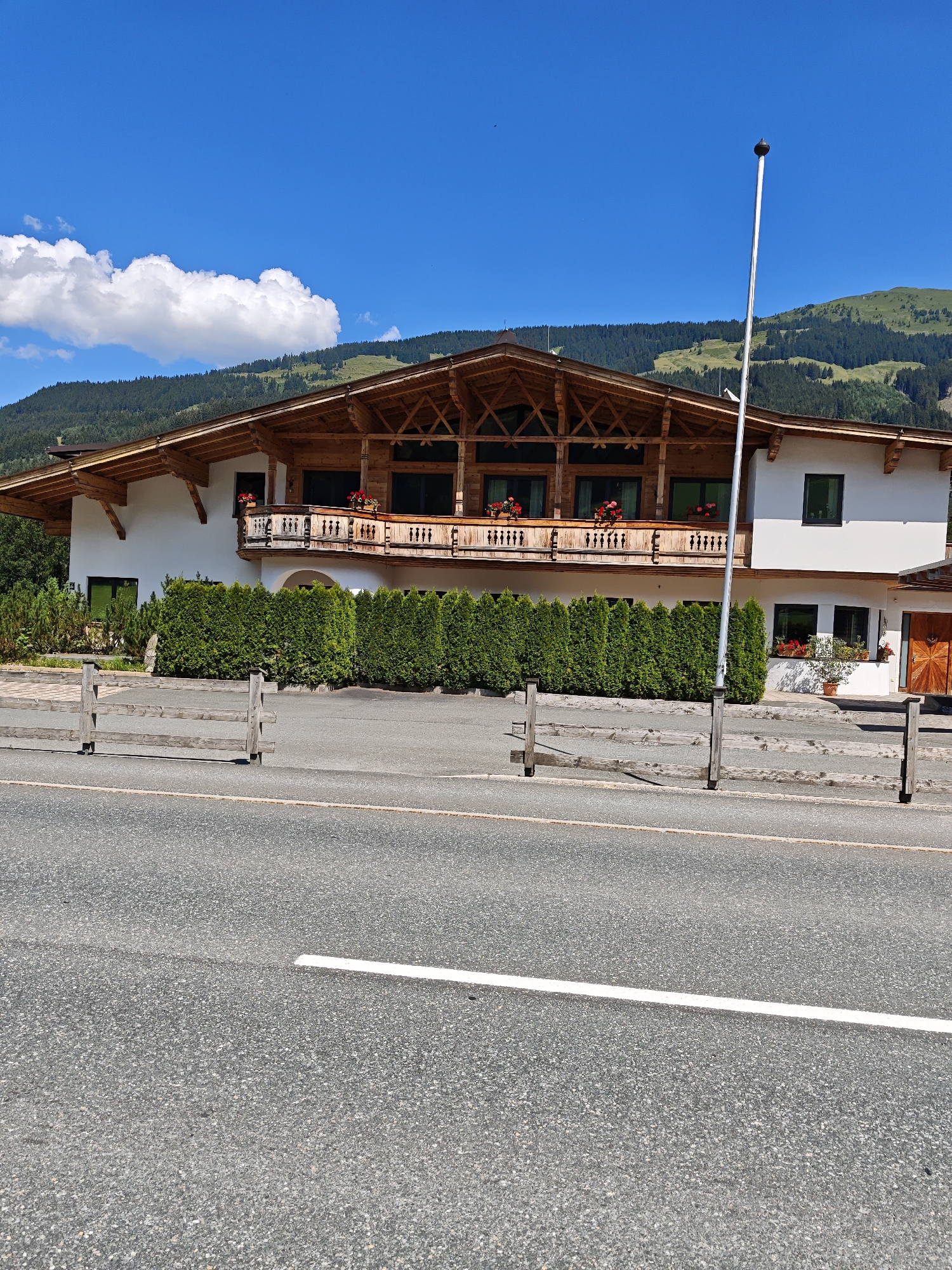 This image shows a traditional Austrian building in Filzen, Austria, on a sunny day. The building is a multi-story structure with a distinctive wooden eaves and a large balcony on the upper level, adorned with flower boxes. The facade is painted white, and there are several dark-framed windows. In front of the building is a neatly trimmed green hedge, and a rustic wooden fence runs along the foreground. A tall, thin pole stands near the center of the image. Beyond the building, rolling green hills and a forest are visible under a bright blue sky with scattered white clouds. The scene appears to be situated near a road, indicated by the asphalt and white line in the foreground. There are no people or visible activities in the image.