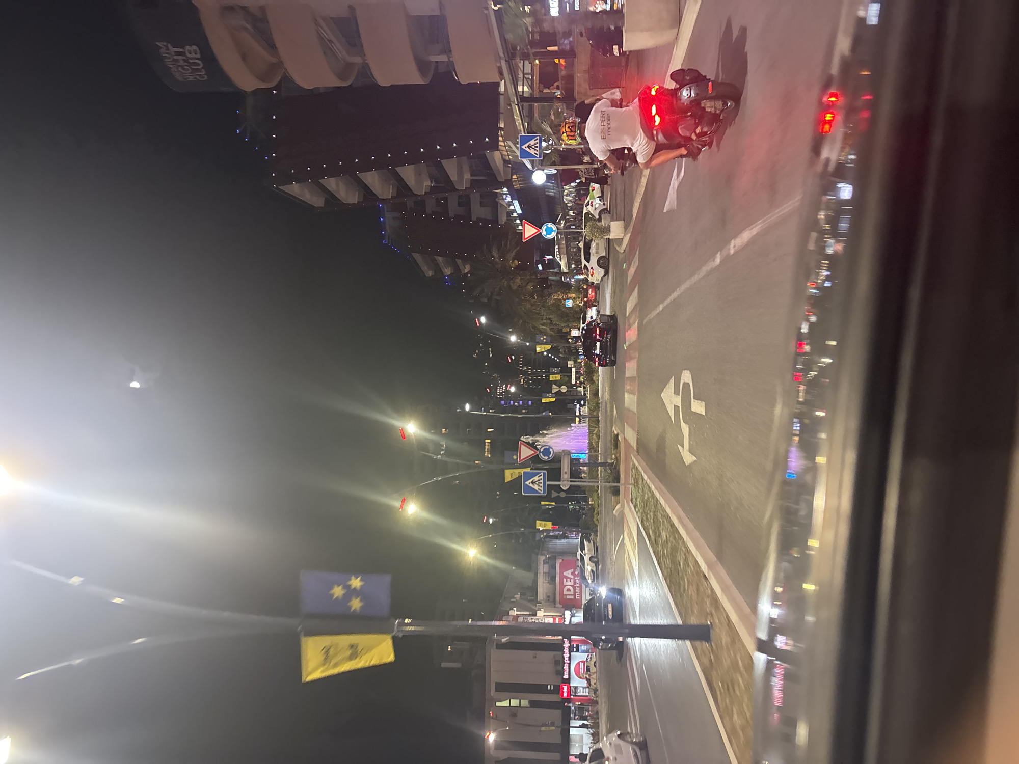 The image captures a street scene at night in Budva, Montenegro. The perspective is from inside a vehicle, looking out through the window.
The primary subject is a person riding a motorcycle, seen from behind. They are wearing a white t-shirt with dark lettering, partially visible as "EXPERT MOBILE". The motorcycle has bright red taillights. The rider is in the right lane of a multi-lane road.
The scene is a city street lined with buildings on the left side, illuminated by streetlights and building lights. One building appears to be a "NIGHT CLUB". Further down the street, there is a fountain with colorful lights, a pedestrian crossing, and various traffic signs, including a roundabout sign and a pedestrian crossing sign. Several cars are visible on the road, some stationary in traffic and others in motion. Yellow and blue flags are hanging from poles along the street. One blue flag features yellow stars, possibly representing the European Union flag.
The time of day is clearly night, indicated by the darkness of the sky and the prevalence of artificial lighting. The street appears wet, suggesting recent rain or damp conditions. The overall atmosphere is lively, with the lights of the city and the presence of traffic indicating an urban environment, likely a popular area. The reflections on the vehicle's window add to the sense of being in a moving vehicle.
Visible text includes "NIGHT CLUB", "EXPERT MOBILE", and "IDEA market". The presence of "IDEA market" suggests a commercial area.