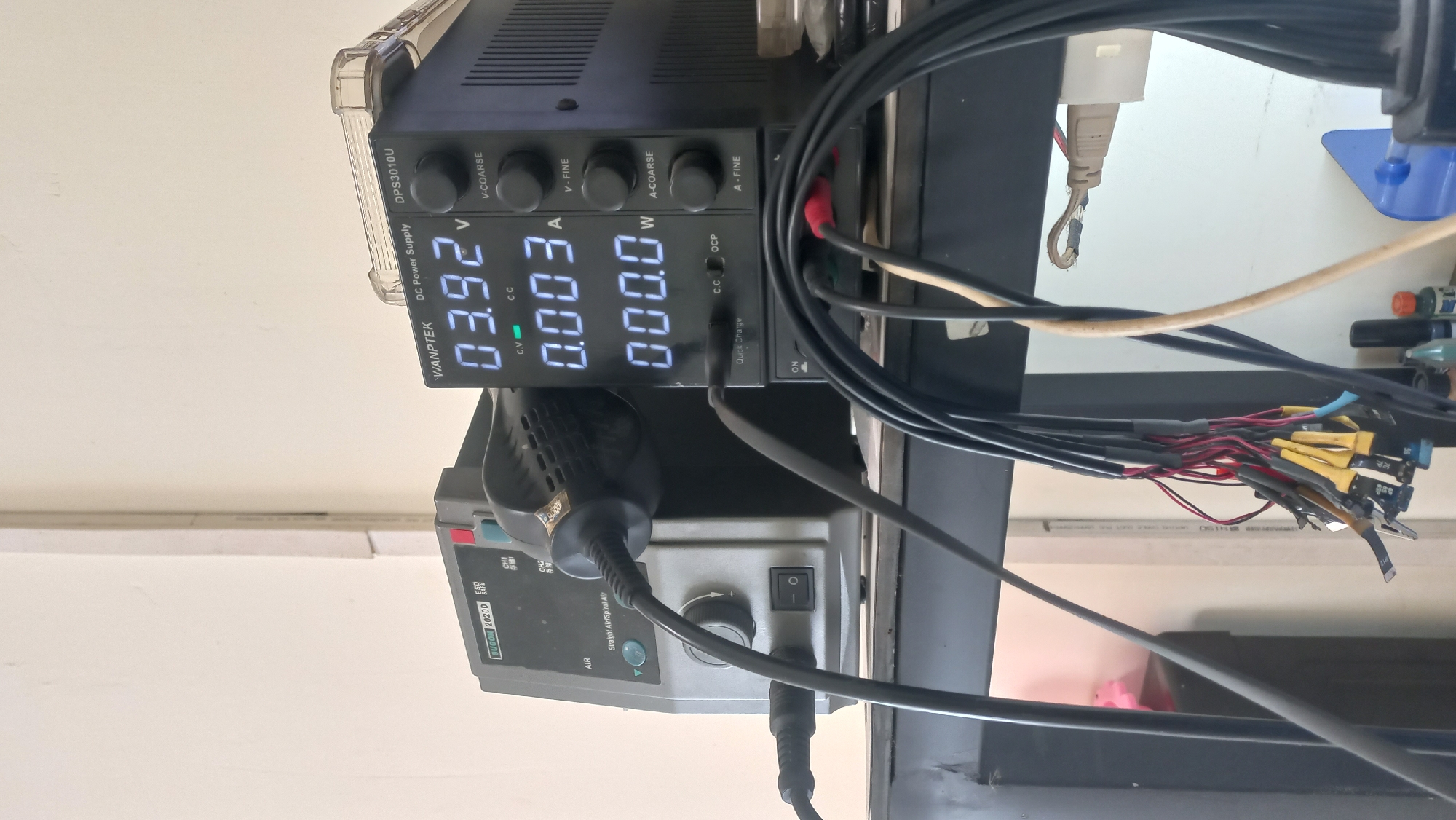 The image shows a close-up of electronic testing equipment on a shelf in what appears to be a workshop or repair area. The equipment includes a gray "SUGON 2020D" hot air rework station with a connected handle, and a black "WANPTEK DC Power Supply DPS3010U". The power supply displays readings of 03.92 V, 0.003 A, and 000.0 W. Several black cables with various connectors hang in front of the equipment, and a beige cable is visible on the right. The background consists of a plain wall with a vertical white conduit. The scene is devoid of people. There is no visible indication of the location being Jetis Indah, Indonesia.