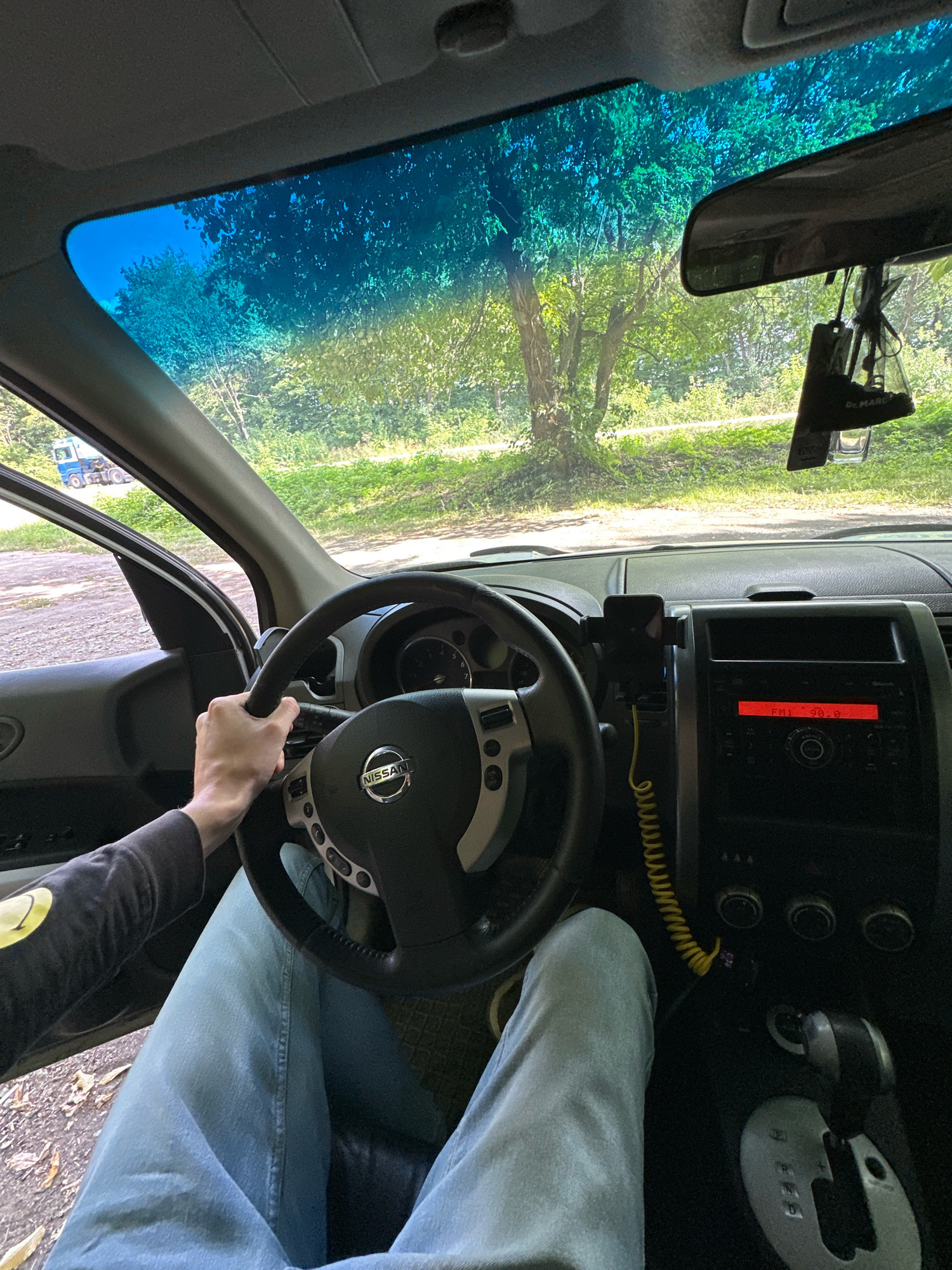 An interior shot from the driver's perspective inside a Nissan vehicle. The driver's hands are on the steering wheel, and their legs, clad in light blue jeans, are visible. The dashboard features a car stereo displaying "FM 90.0" and a phone mount. Outside the car, visible through the windshield, is a tree-lined area with a dirt road and a blue and white truck in the distance. The scene appears to be located near Shatava, Ukraine, given the context.