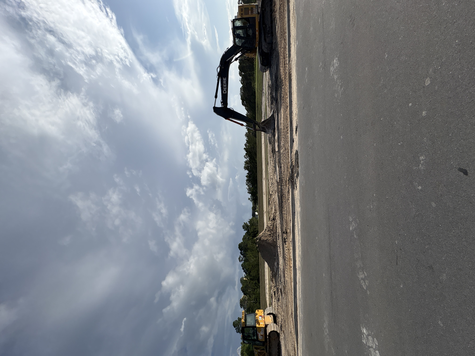 The image depicts a construction site in Panama City Beach, United States of America. A yellow John Deere excavator is actively digging into a dirt pile beside a paved road. Another yellow bulldozer is visible in the lower part of the image. The background consists of trees and a cloudy sky.
