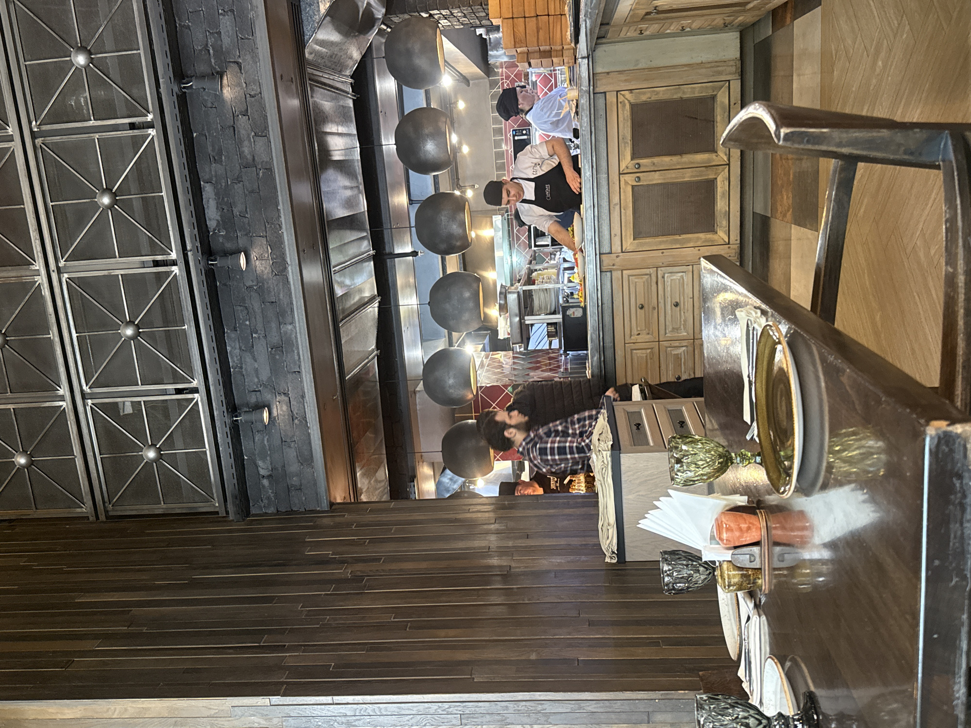 The image shows the interior of a restaurant in Yerevan, Armenia. Two chefs are working in the kitchen. A patron is seated at a table in the foreground. The restaurant features dark wood flooring and a metal decorative partition on the left side. Large spherical pendant lights hang above the kitchen area. The table is set with plates, glasses, and napkins.