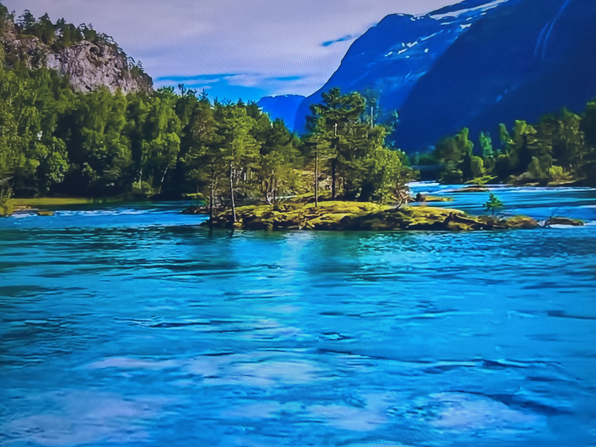 The image depicts a vibrant, natural landscape. In the foreground, a clear, turquoise river flows rapidly, exhibiting subtle ripples and currents. The water reflects the colors of the sky and surrounding environment. In the middle ground, a small, verdant island is populated with trees, with a mountain looming in the background. Other trees also are on the left side. The sky above is a mix of white and blue, with a hint of clouds. No people or animals are visible in the scene. The location is not in the city of Basingstoke and Deane, United Kingdom. This is a landscape image of a river in a forested area, likely somewhere in the mountains of Scandinavia.