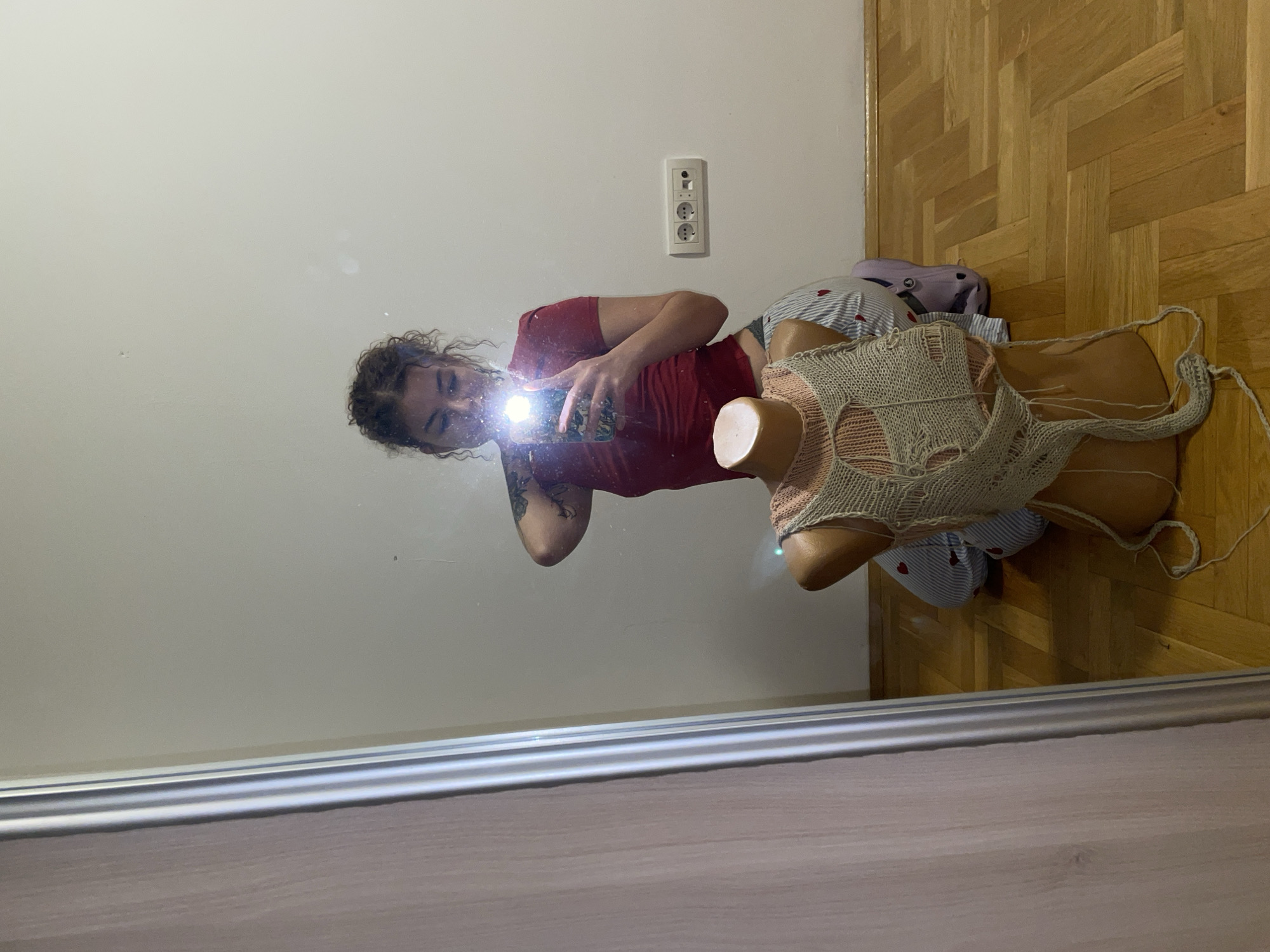 The image depicts an indoor scene in Boreti, Montenegro, captured through a mirror reflection. A female individual with curly dark hair and a visible tattoo on her left arm is kneeling on a wooden herringbone parquet floor. She wears a red short-sleeved top and striped pants patterned with red hearts. The individual is actively taking a mirror selfie, holding a smartphone with both hands, its flash illuminated and directed towards her face. In the foreground, positioned between the camera and the reflected individual, is a headless, skin-toned female mannequin torso. A distressed, light-colored knitted garment, featuring holes and loose threads, is draped over the mannequin, revealing a pink knitted layer beneath it. The background consists of a plain white wall. A white electrical outlet unit, containing two power sockets and a data port, is affixed to the wall to the right of the reflected individual. A wooden frame is visible on the left side of the mirror. A partial purple object is discernible on the floor behind the mannequin.