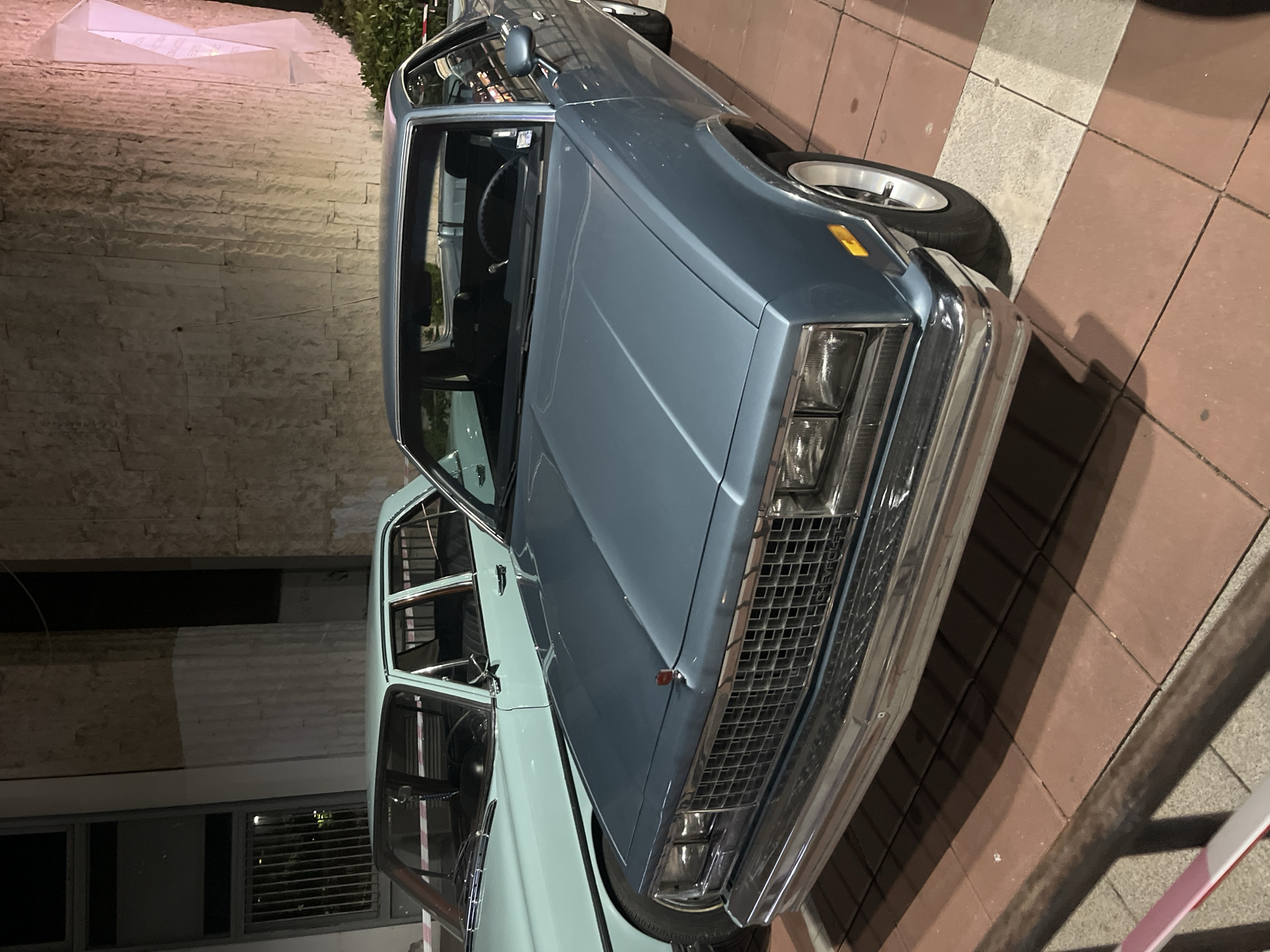The image shows two classic cars parked on a brick and concrete surface. The car in the foreground is a blue coupe with a prominent chrome grille and rectangular headlights. The other car, a light green sedan, is situated partially behind the coupe. The setting appears to be a city street, with a stone building visible in the background.