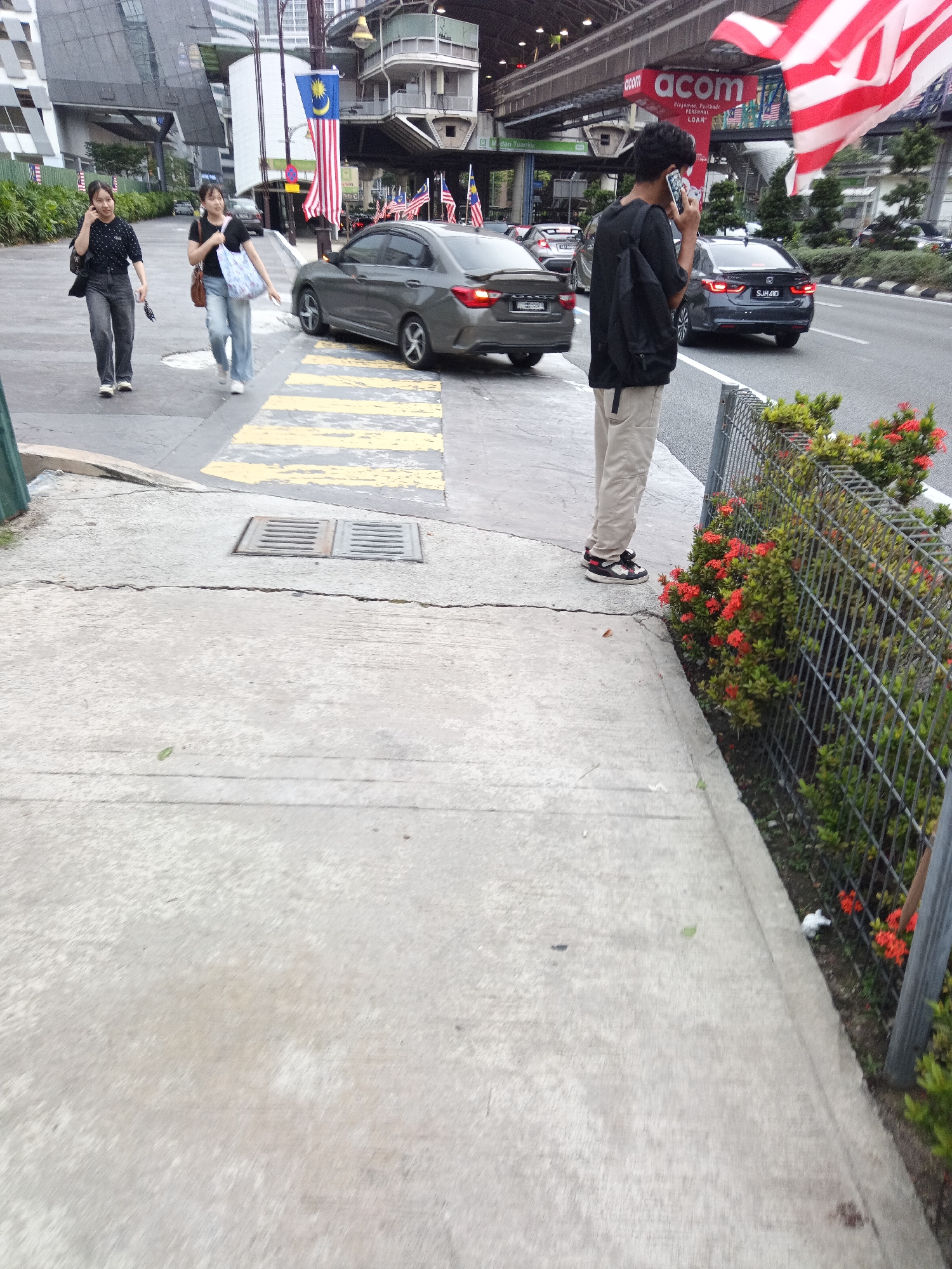 The image depicts a street scene in Kuala Lumpur, Malaysia, likely near a commercial or transportation hub given the presence of a pedestrian overpass and advertisements. Two young women are walking on a sidewalk, one talking on her phone and carrying a bag. A young man stands on the sidewalk facing the street, speaking on his phone and wearing a backpack. He is dressed in a dark t-shirt and light-colored pants. Several cars are on the road, including a dark-colored sedan in the foreground and another similar car further back. The Malaysian flag is prominently displayed, waving in the breeze, along with other flags. The environment includes buildings, trees, a metal fence with flowering plants, and a street grate on the sidewalk. The time of day appears to be daytime.