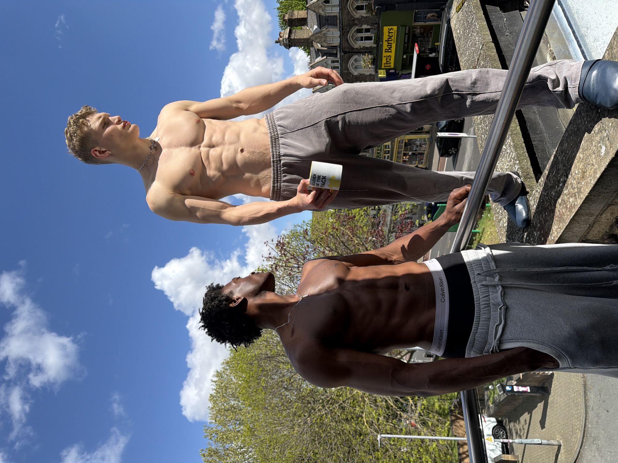 Two men, viewed from a low angle against a blue sky with scattered clouds, stand outdoors. The man at the top, fair-skinned with blonde hair, is shirtless, wearing grey sweatpants and blue shoes. He holds a white and yellow mug in his left hand. The other man, who has dark skin, is also shirtless, wearing grey sweatpants with a visible Calvin Klein waistband. Both men are looking upwards. In the background, a building with a sign that reads "Ibiza Barbers" is visible, suggesting a location in London. A metallic railing is positioned in the foreground, partially obscuring the lower portions of both men. Green trees are also visible in the background.