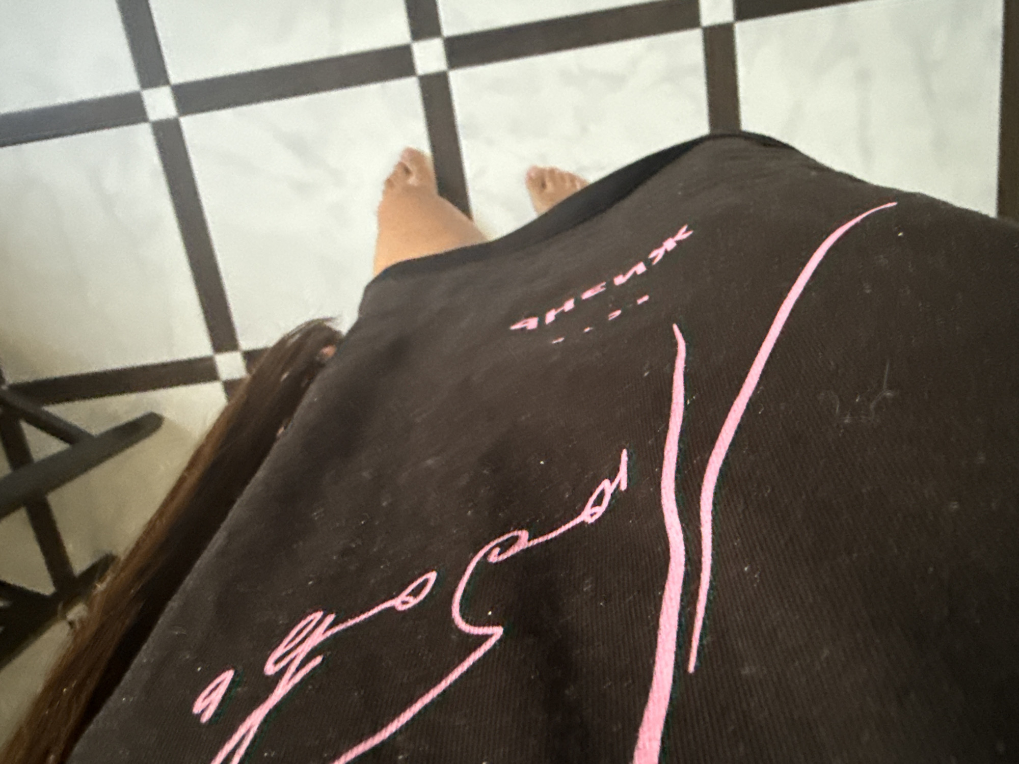 The image shows a close-up of a person's legs and lower torso, wearing a dark garment with pink graphic designs. The garment appears to be made of a fabric with a visible texture, possibly cotton or a blend. The pink design is abstract and flowing, with some elements resembling handwriting or brushstrokes. A few small white specks, possibly dust or lint, are visible on the garment.
The person's legs are bare below the garment. The right leg is bent at the knee, and the feet are visible. The toes are distinct, and the skin tone suggests a Caucasian individual. The floor beneath the person is tiled with a black and white checkered pattern, creating a stark contrast with the dark garment and skin. The lighting in the scene appears to be artificial, casting soft shadows.
The background shows a portion of what appears to be furniture with dark legs, possibly a chair or table, suggesting an indoor setting. The overall composition is slightly tilted, giving a dynamic feel to the image.
Without further information or context, it is impossible to determine the specific location within the city of Chișinău, Moldova. The image lacks any identifiable landmarks, signage, or architectural details that would pinpoint its exact geographical setting.