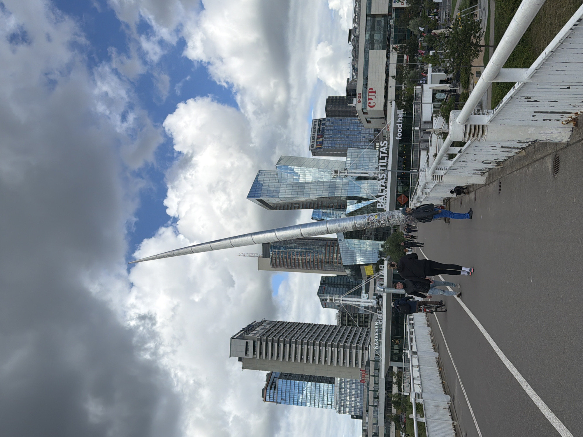 This image captures a wide-angle view of a bridge in Vilnius, Lithuania, with the modern cityscape in the background. The bridge features a pedestrian walkway and a bike lane, separated by white lines. Several people are visible on the bridge; two individuals are walking towards the camera, one on a bicycle, and another person is walking a dog. A tall, sleek, silver sculpture rises from the center of the bridge, reaching towards a cloudy sky with patches of blue.
In the background, a cluster of contemporary buildings with glass facades dominate the skyline. Prominent signage on these buildings includes "BALTAS TILTAS" (White Bridge) and "CUP," a shopping center. The overall environment appears to be an urban setting, possibly a business or commercial district, with trees and green spaces visible to the right of the bridge. The lighting suggests it is daytime, with diffused sunlight filtering through the clouds.