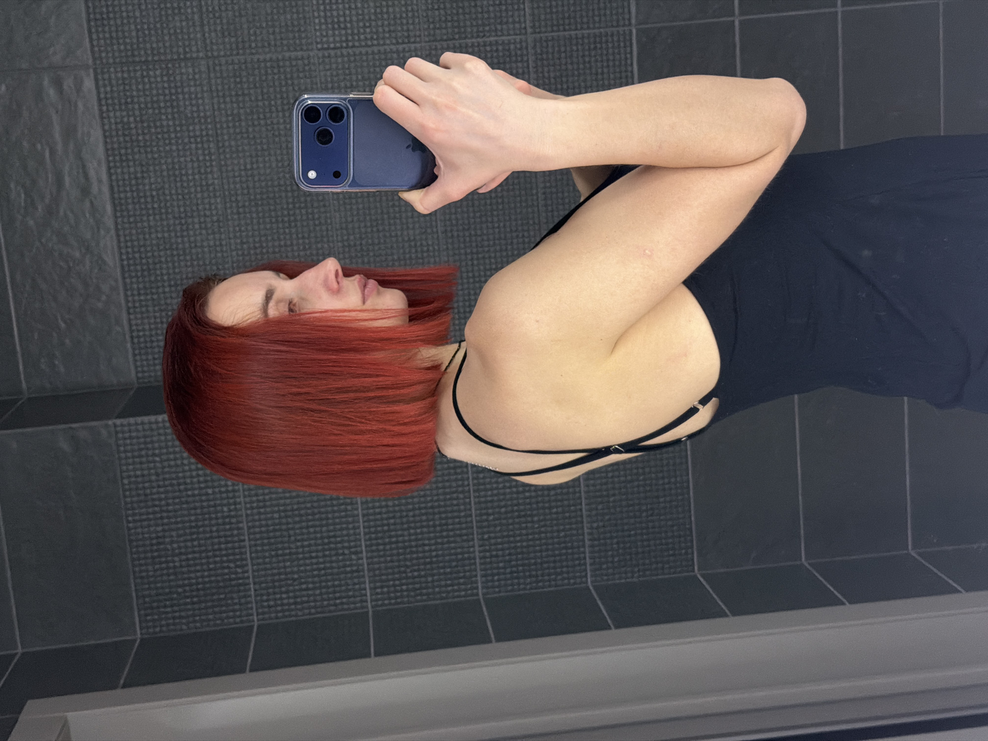 A fair-skinned woman with short, red hair is taking a selfie with a blue smartphone in a bathroom. She is wearing a black top with spaghetti straps. The background consists of dark gray tiled walls. The location may be Chaikovsky, Russia.