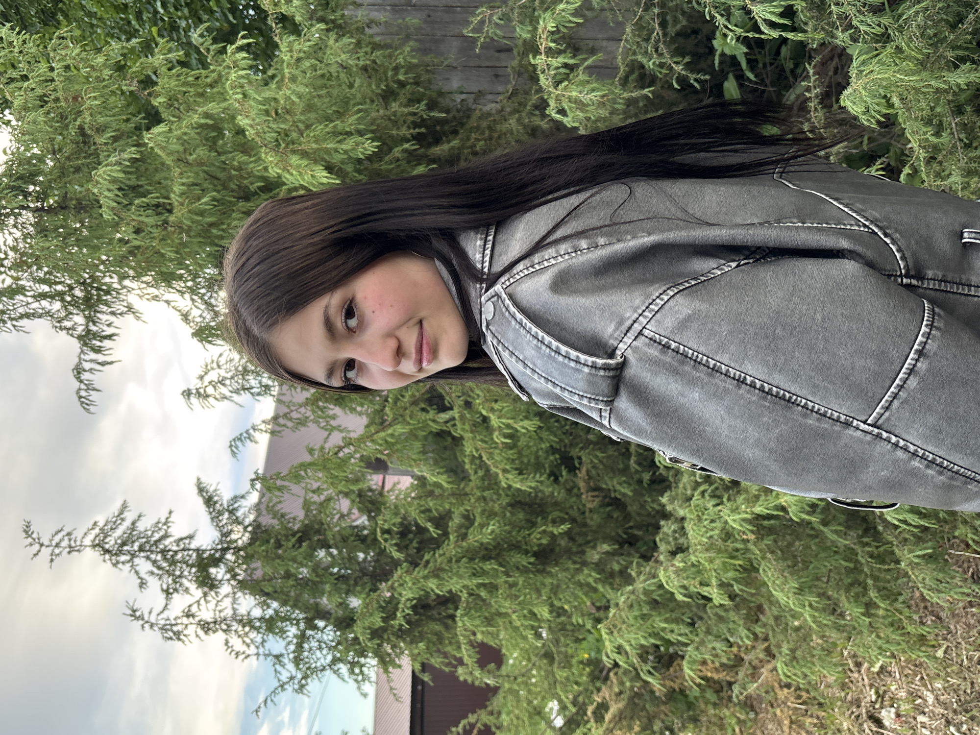 A young person stands outdoors, likely in a yard or garden. They have long, dark hair and are wearing a grey denim jacket. Lush green bushes and trees fill the background, with a wooden fence partially visible at the top. The lower portion of a building with a brown roof is also visible, suggesting a residential area. The sky is cloudy. Based on the location provided, the scene appears to be in Фоки, Russia.