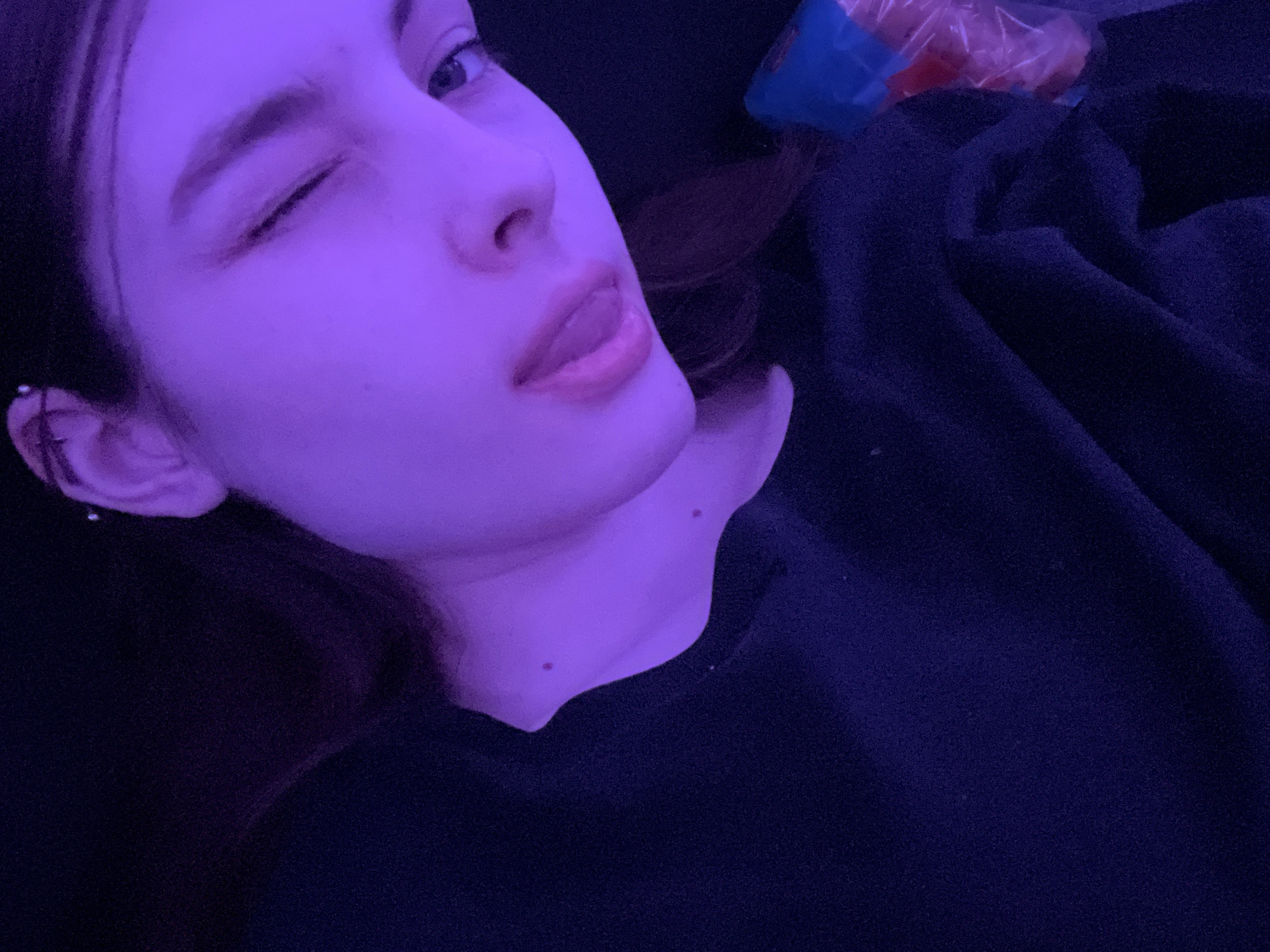 A young person with fair skin and dark hair is lying down, winking one eye and sticking out their tongue slightly. They are wearing a dark-colored top. Their ear has multiple piercings. Two small moles are visible on their neck. The image appears to be taken indoors under purple lighting. A plastic bag with contents is partially visible in the background. There is no indication of the location being Saint Petersburg, Russia.