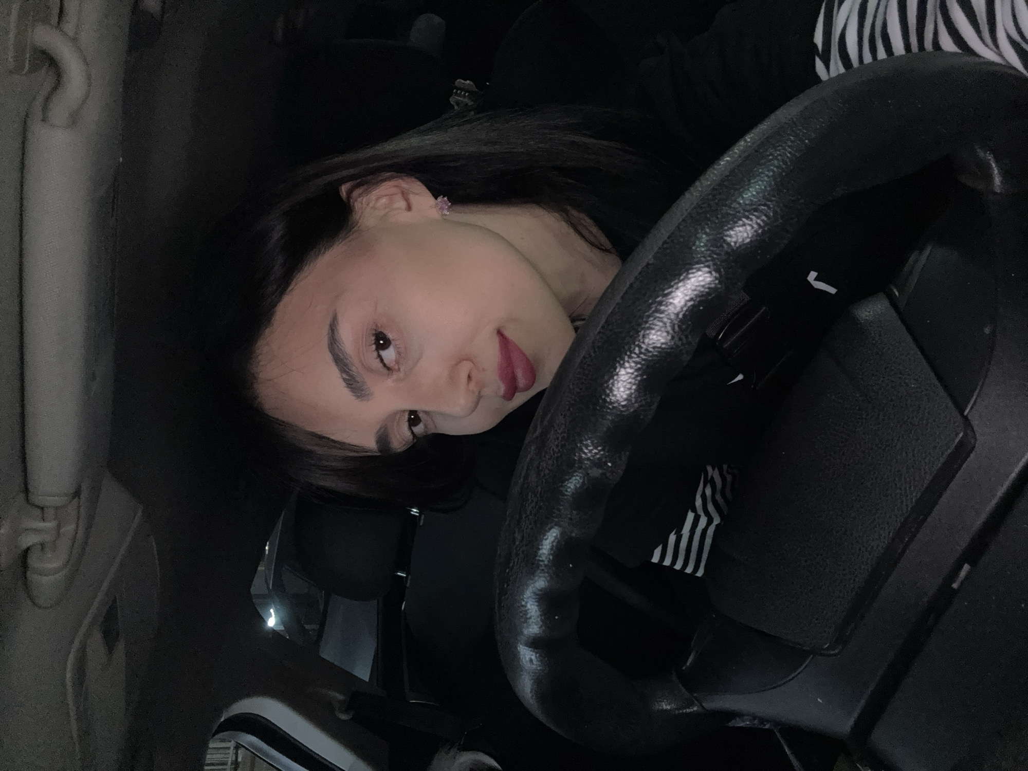 A young woman is captured in a close-up, rotated 90 degrees clockwise, inside a car in Tbilisi, Georgia. She is positioned with her head tilted, looking directly at the camera with a playful expression, her tongue slightly out. Her long, dark hair frames her face, which features dark eyebrows, eyeliner, and dark lipstick. She wears a dark top, with a black and white striped sleeve visible on the right side of the frame. A small pink stud earring is noticeable in her left ear.
The foreground is dominated by the woman's upper body and a prominent black steering wheel, which has a textured cover. A distinct white logo, identifiable as a Nike swoosh, is visible on this steering wheel cover. The car's interior appears dark and dimly lit, suggesting the photograph was taken at night or in a low-light environment. Portions of the car's headliner or interior pillar are visible on the left side, further confirming the setting. The image conveys a casual and personal moment.
