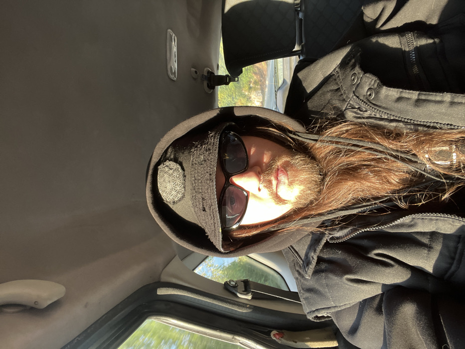 A single individual, identified as an adult male, is captured in a front-facing photograph from within a vehicle's interior. The subject has long, medium-brown hair and a full beard, wears black oval-shaped sunglasses, a black hooded sweatshirt with the hood up, and a black bucket hat. The hat features a prominent circular patch on its front with a geometric, patterned design. Over the sweatshirt, a black sleeveless vest or jacket with a central zipper and multiple snap-closure pockets is worn. Strong direct sunlight enters the vehicle from the left, illuminating the left side of the subject's face and casting reflections on the sunglasses. The vehicle's interior shows a light-colored headliner, a grab handle on the left, and a black headrest of an adjacent seat on the right. Through the windows, a blurred outdoor environment with green foliage is visible, indicating the vehicle is in motion or the background is out of focus. The specific location in the city of Rijeka Crnojevića, Montenegro, cannot be determined from the visual evidence present in the image.