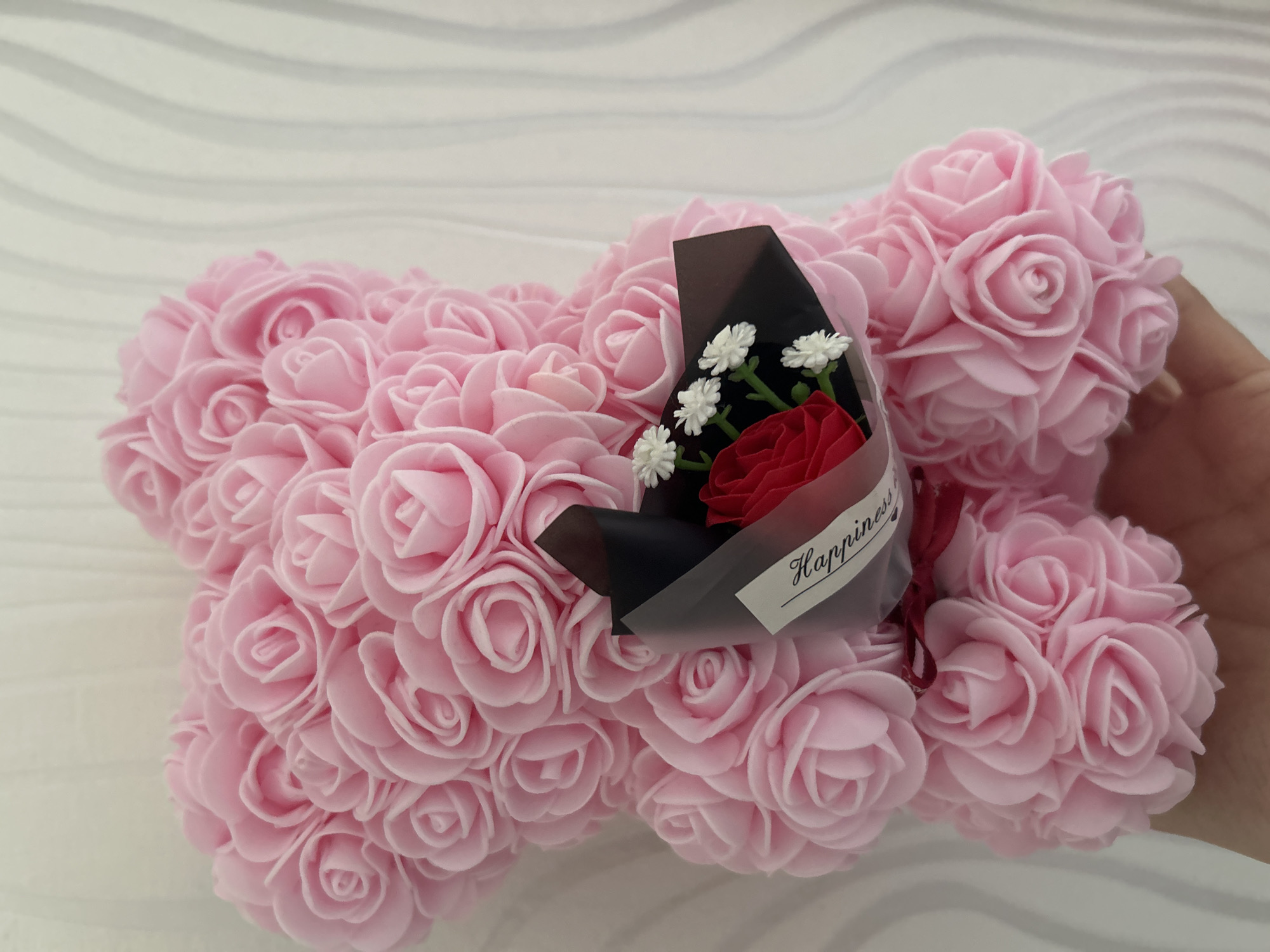 The image shows a foam rose bear with a small bouquet. The bear is composed of numerous pink foam roses, creating a dense, textured surface. A small bouquet rests on the bear, featuring a red rose and a few small white flowers, wrapped in black paper with a "Happiness" tag. A hand is partially visible on the right side of the image. The background is a white wooden surface. There is no evidence to suggest this picture was taken in Strasbourg, France.