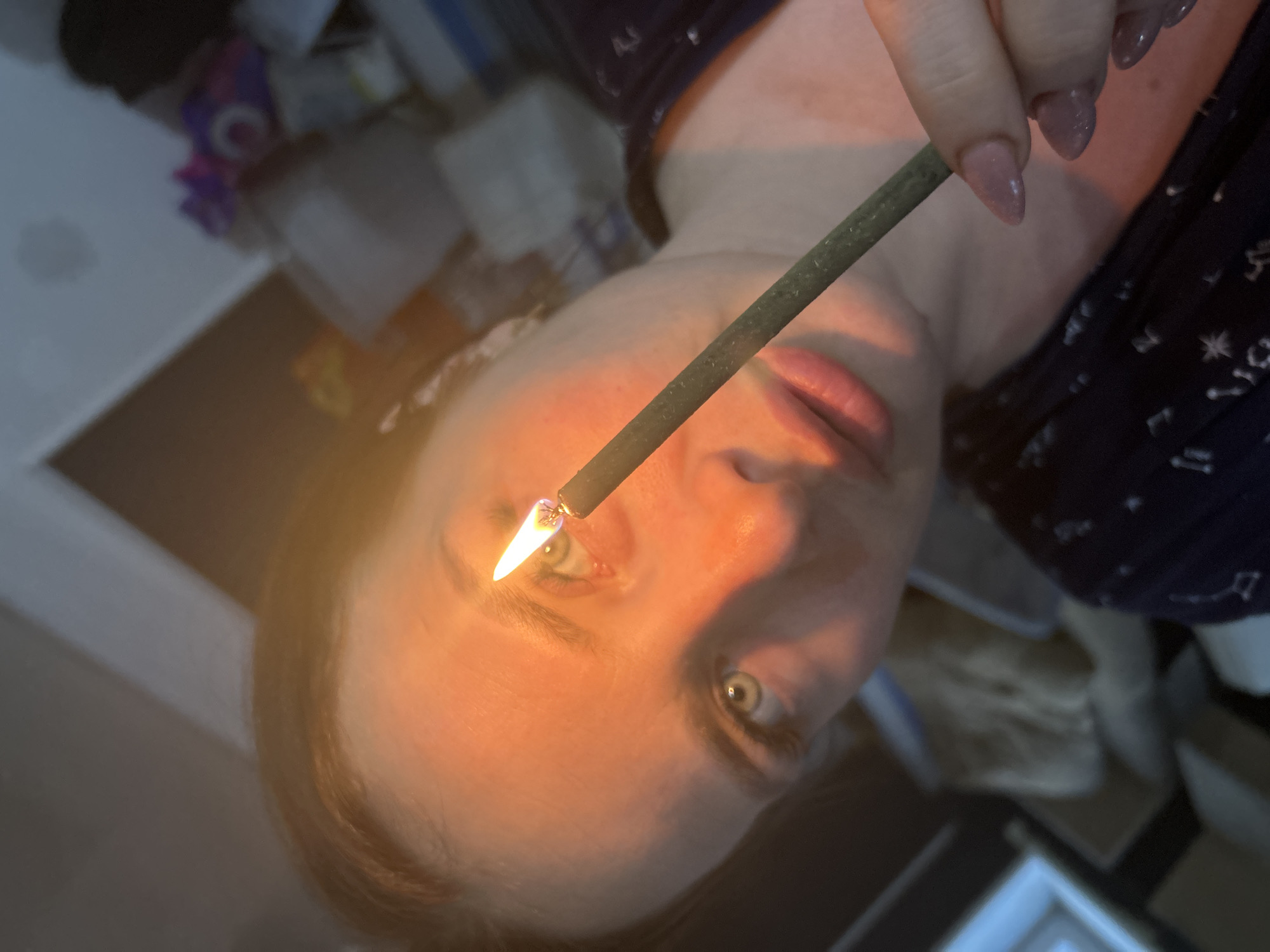 A person is holding a lit incense stick close to their face, appearing to touch their forehead. The person has light skin, light eyes, and light hair. They are wearing a dark-colored top with a constellation pattern. The background includes blurred objects and surfaces. The location is unknown, and there is no indication it is Rusalevka, Russia.