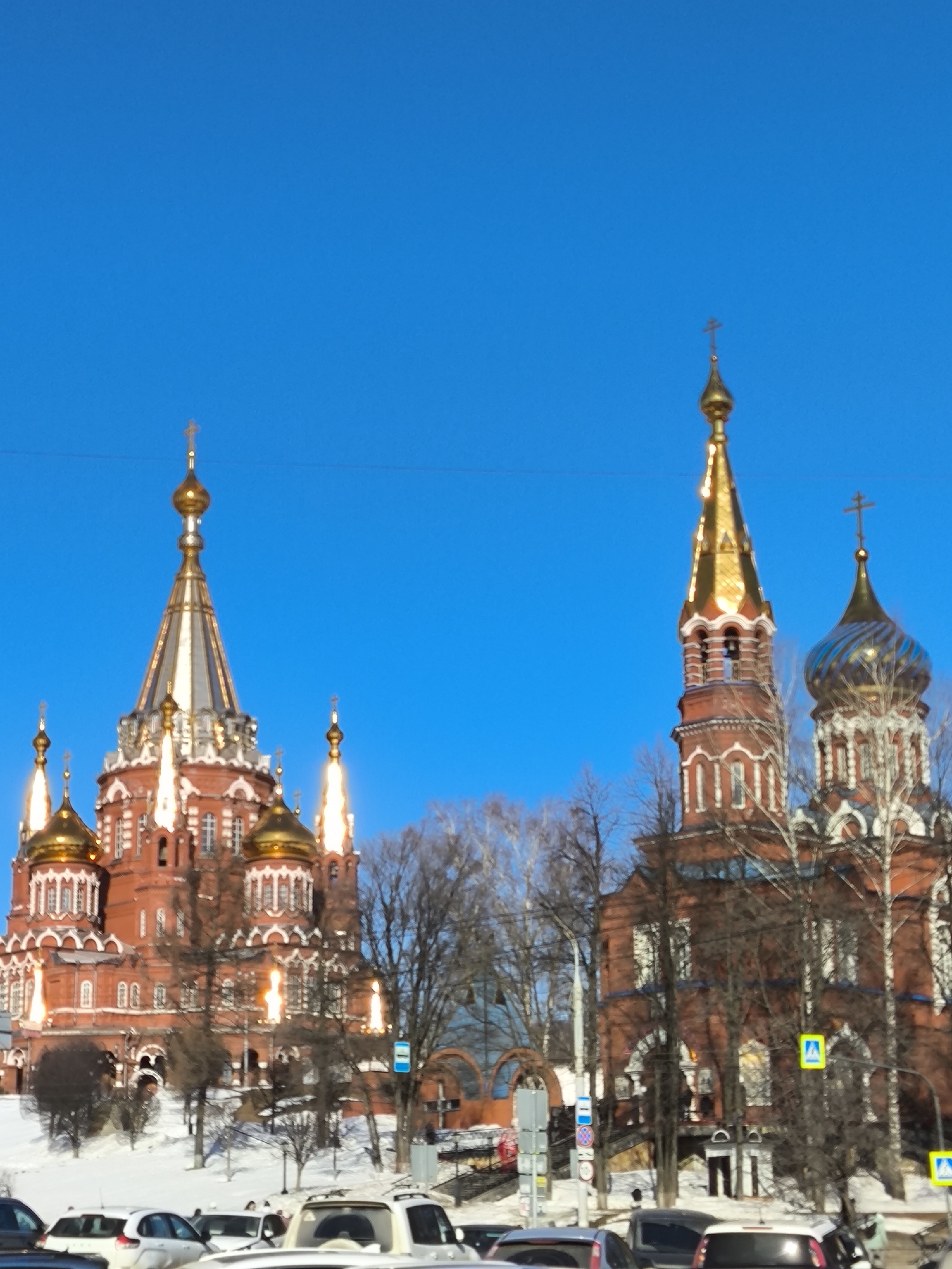 The image displays a winter scene in Izhevsk, Russia, featuring two prominent red-brick Orthodox cathedrals under a clear, bright blue sky. The ground in the mid-ground and foreground is covered in snow.
On the left, the Cathedral of St. Michael stands on a snow-covered hill. This large, multi-domed structure is constructed of red brick with white architectural detailing. It features a central tall, conical spire topped with a golden cross, flanked by several smaller golden onion domes and four additional spires with golden crowns and crosses. The roof of the main spire is silver-colored, and the lower domes are golden.
To the right, partially obscured by bare deciduous trees, is the Alexander Nevsky Cathedral. This structure is also red brick with white trim. It features a prominent golden-domed bell tower (belfry) on the left, topped with a golden cross. To its right, another section of the cathedral has a blue and gold patterned onion dome, also topped with a golden cross. The trees in the mid-ground are sparse, indicating winter, with branches devoid of foliage.
The foreground shows a street level with multiple parked and possibly moving automobiles of various makes and colors, including white, silver, and black vehicles. Several street signs are visible, including a blue and white bus stop sign and yellow square signs with a blue pedestrian crossing symbol. Power lines or utility cables are visible across the upper portion of the frame. No discernible individuals are performing specific actions. The overall environment suggests a cold, clear, sunny winter day.